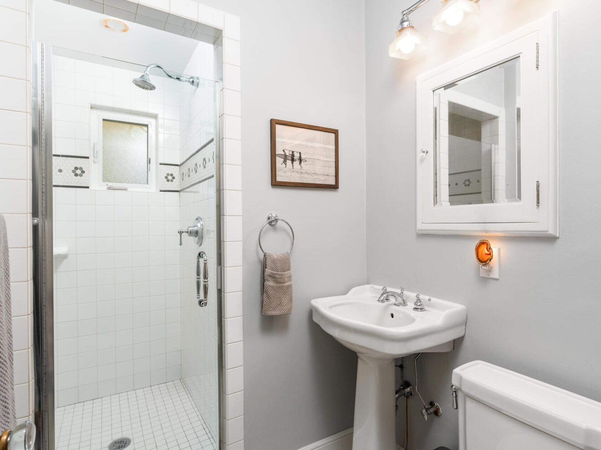 A bathroom with a white tiled shower featuring a glass door and floral accents. The vanity includes a white sink with a mirror above, flanked by a towel ring with a beige towel. A framed picture hangs on the wall above the towel.