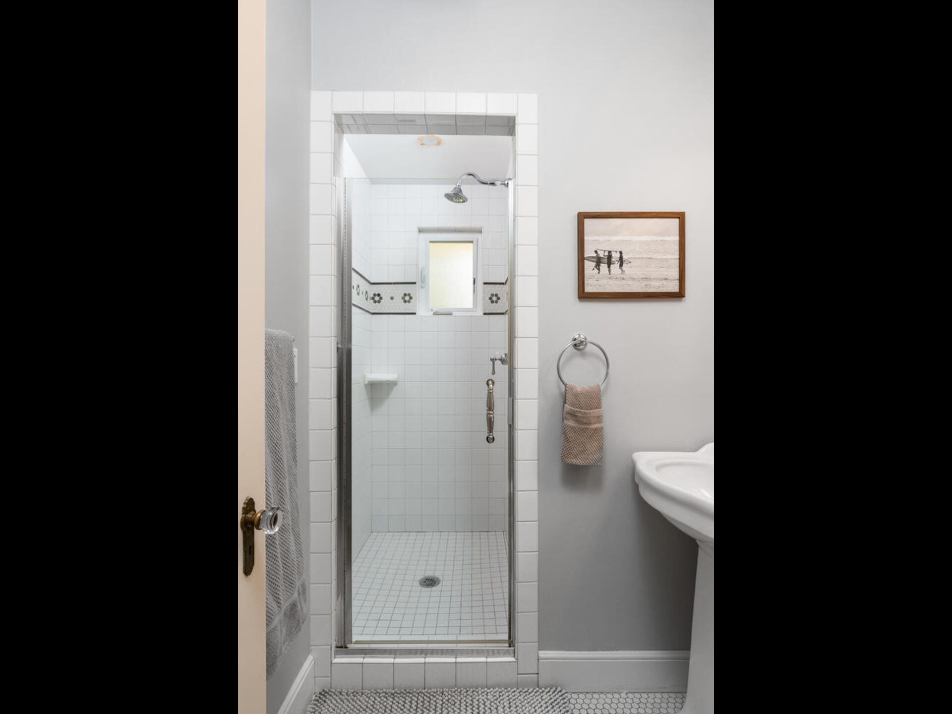 A small, tiled shower with a glass door and a white tile design featuring black accents. A towel hangs on a ring beside a white pedestal sink. The wall above the sink has a framed picture. The floor is covered with hexagonal tiles.
