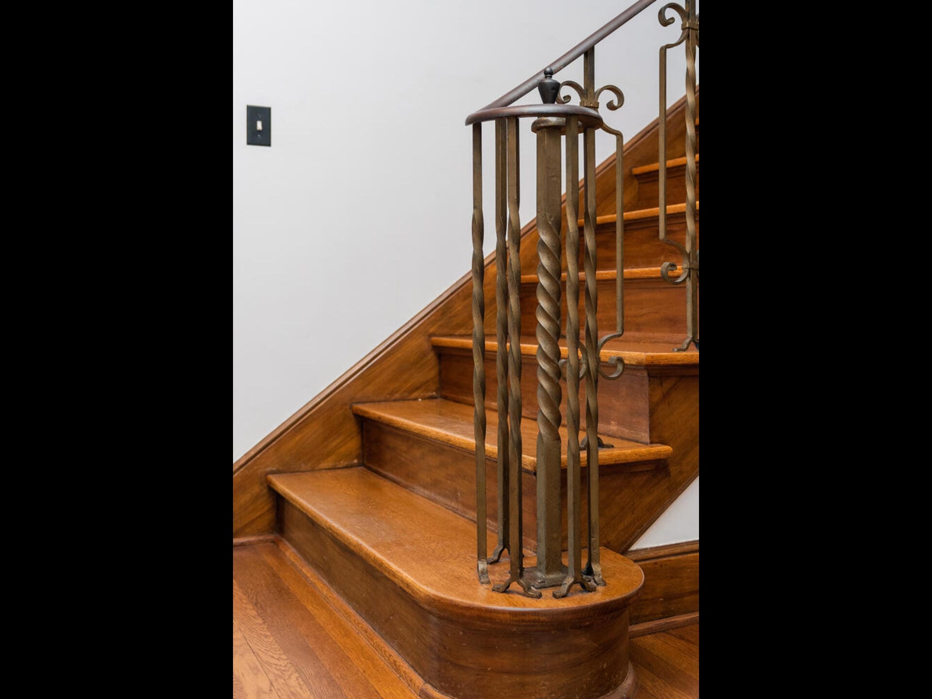 A wooden staircase with a metal handrail features twisted metal balusters. The steps have a warm, polished finish, and the staircase is against a white wall with a light switch visible.
