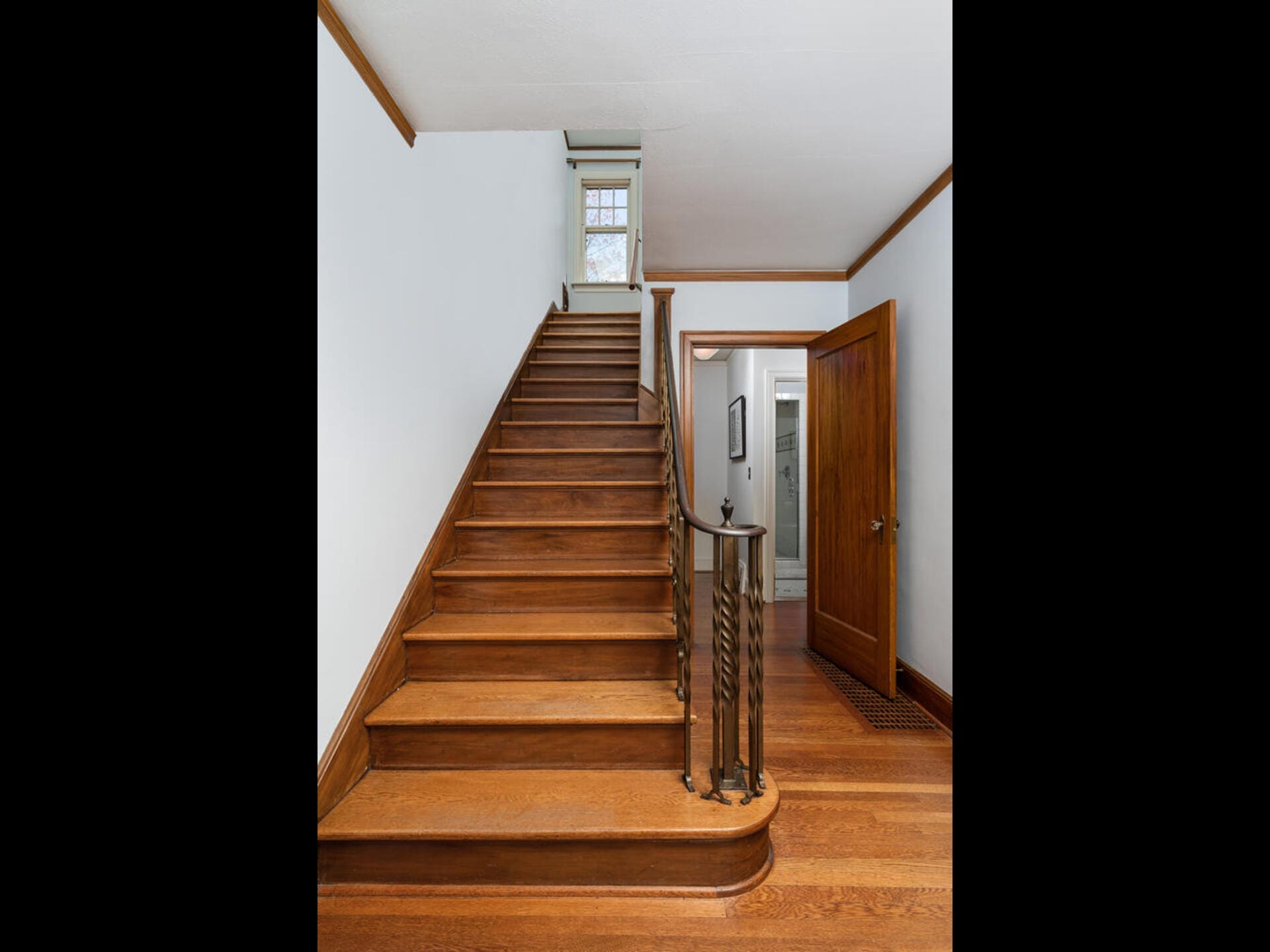 A wooden staircase with brown handrails leads to an upper floor. Theres a small window at the top. On the right, an open brown door reveals a hallway with light blue walls and framed artworks. The floor is wooden.