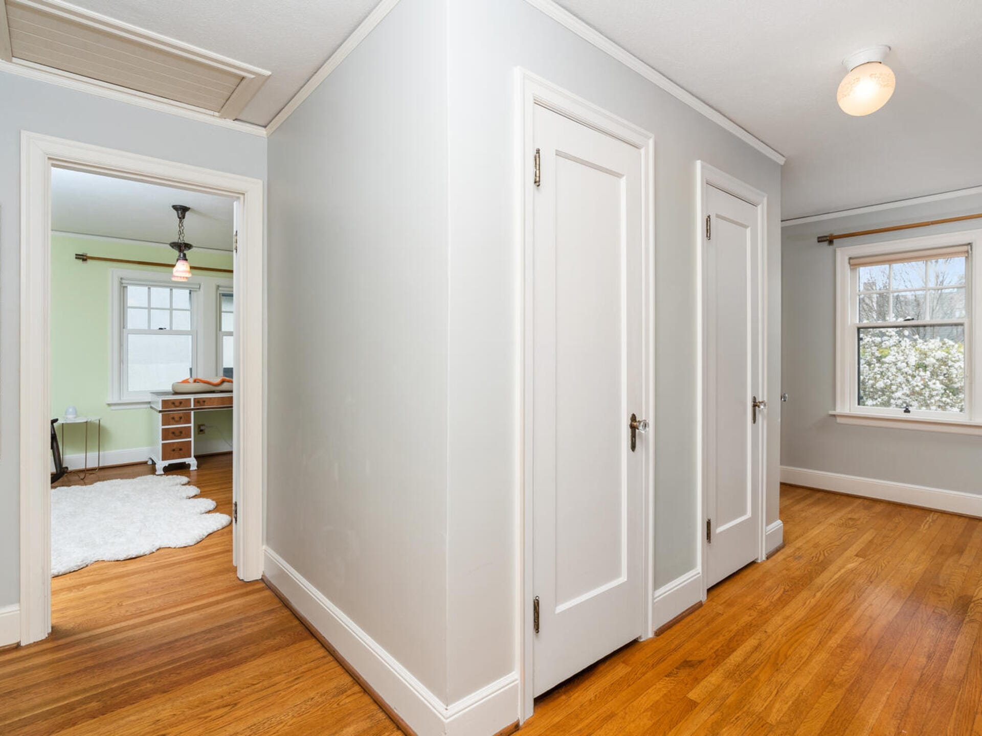 A hallway with light wood flooring and two closed white doors. An open doorway reveals a room with a desk, chair, lamp, and a large white rug. Another door is open to a room with a window displaying an outdoor view. Walls are painted light grey.