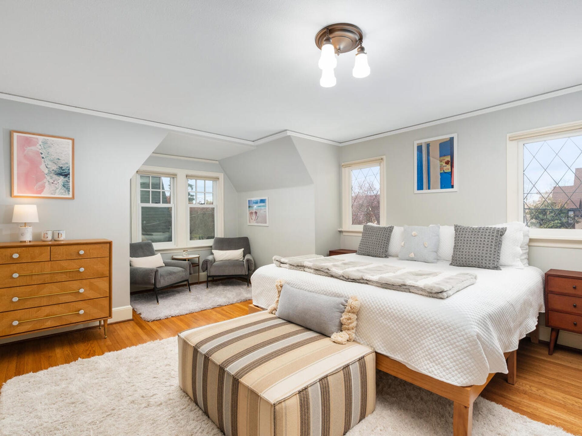 A cozy bedroom with a large bed, gray walls, and wooden floors. Two armchairs sit by the window. A striped ottoman is at the foot of the bed. Artwork hangs on the walls, and a wooden dresser is in the corner. Warm lighting adds comfort.