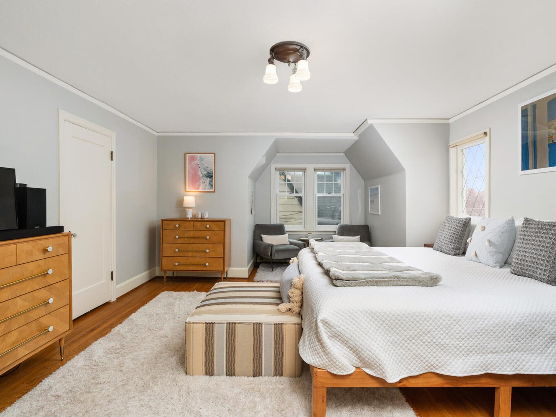 A bright bedroom features a large bed with decorative pillows and a striped bench at its foot. Light gray walls, two small armchairs by a window, a dresser with a lamp and art above it, and a plush rug complete the cozy space.