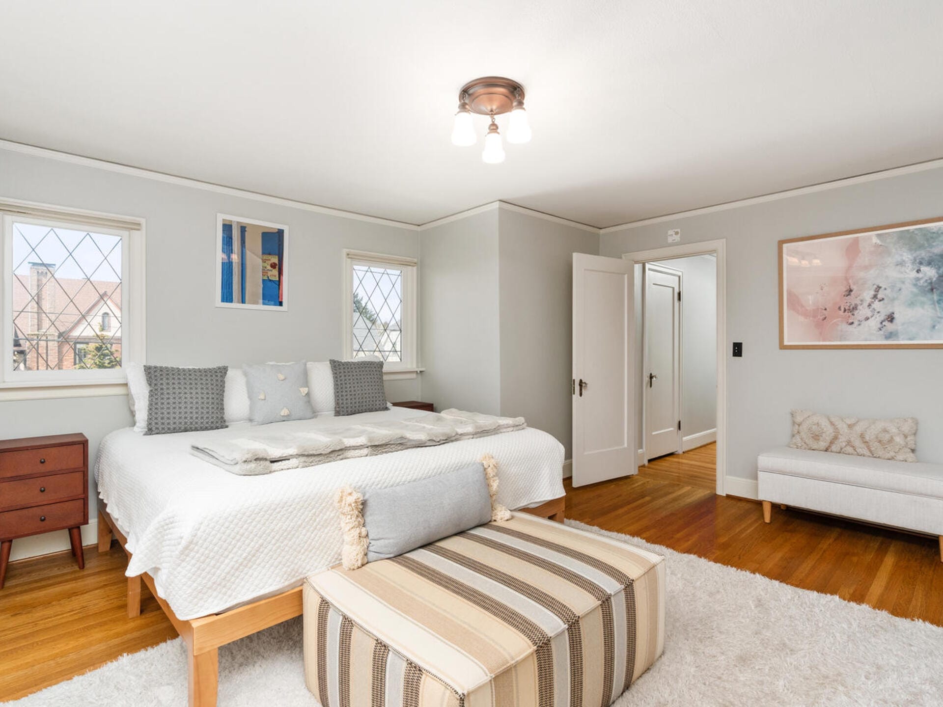 A bright bedroom with a large bed, patterned pillows, and a throw. A striped ottoman sits at the foot of the bed. The room features hardwood floors, light walls, a painting, a small bench, bedside table, and windows with a view.
