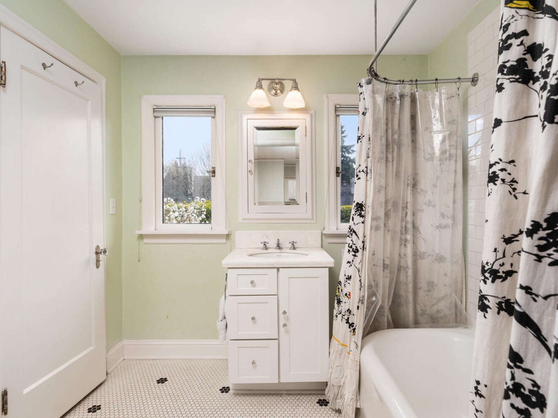 A bright bathroom with green walls and a white vanity with a mirror. Two windows provide natural light. A bathtub with a floral-patterned shower curtain is on the right, and the floor has small black and white tiles.