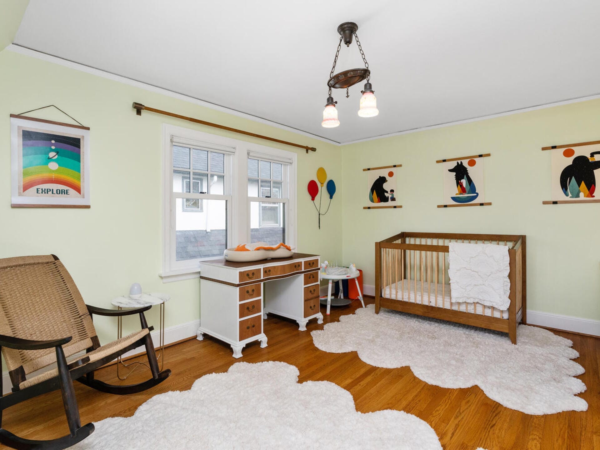 A cozy nursery with light green walls features a wooden crib, a vintage-style dresser with a changing pad, a rocking chair, and whimsical wall art with colorful balloons. Fluffy white rugs cover the hardwood floor beneath a classic ceiling light.