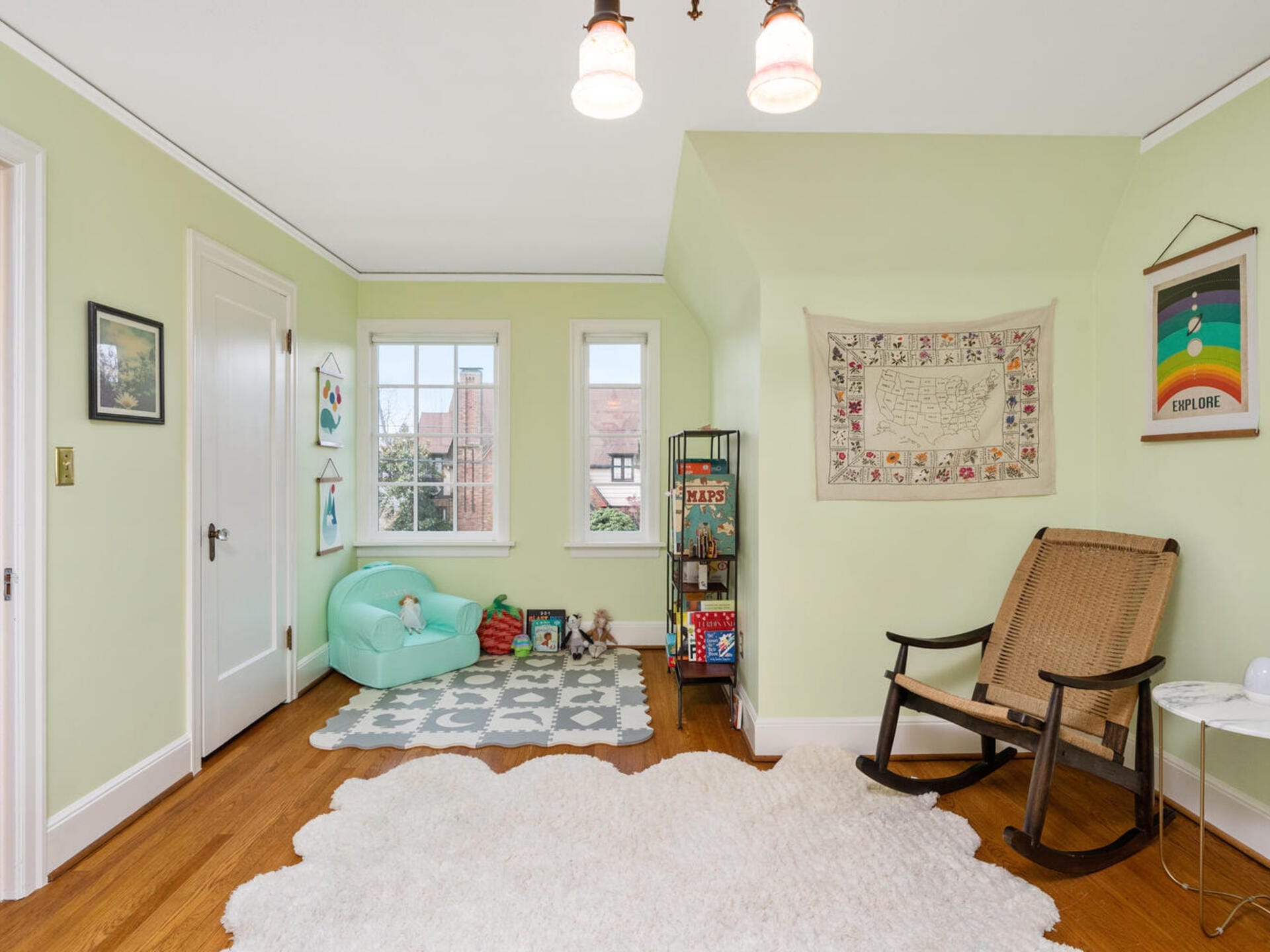 A cozy childrens room with light green walls, a soft shag rug, a comfortable green bean bag chair, and a wooden rocking chair. Shelves hold childrens books and toys, while colorful artwork decorates the walls. Natural light streams through the windows.