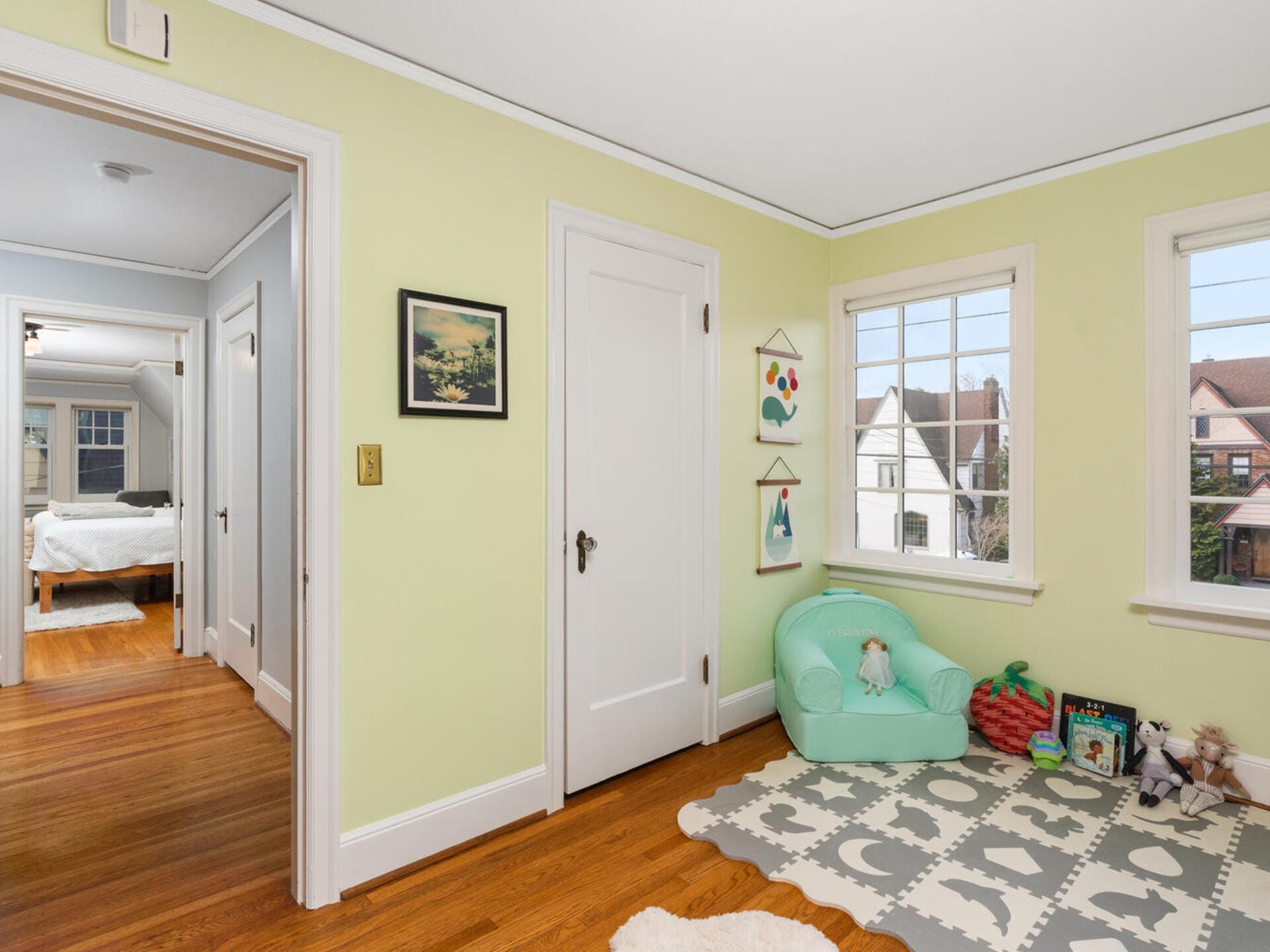 A childs playroom with pastel green walls, a small green armchair, soft play mats on the wooden floor, and toys including books and stuffed animals. Windows provide a view of houses outside, and a hallway leads to a bedroom.