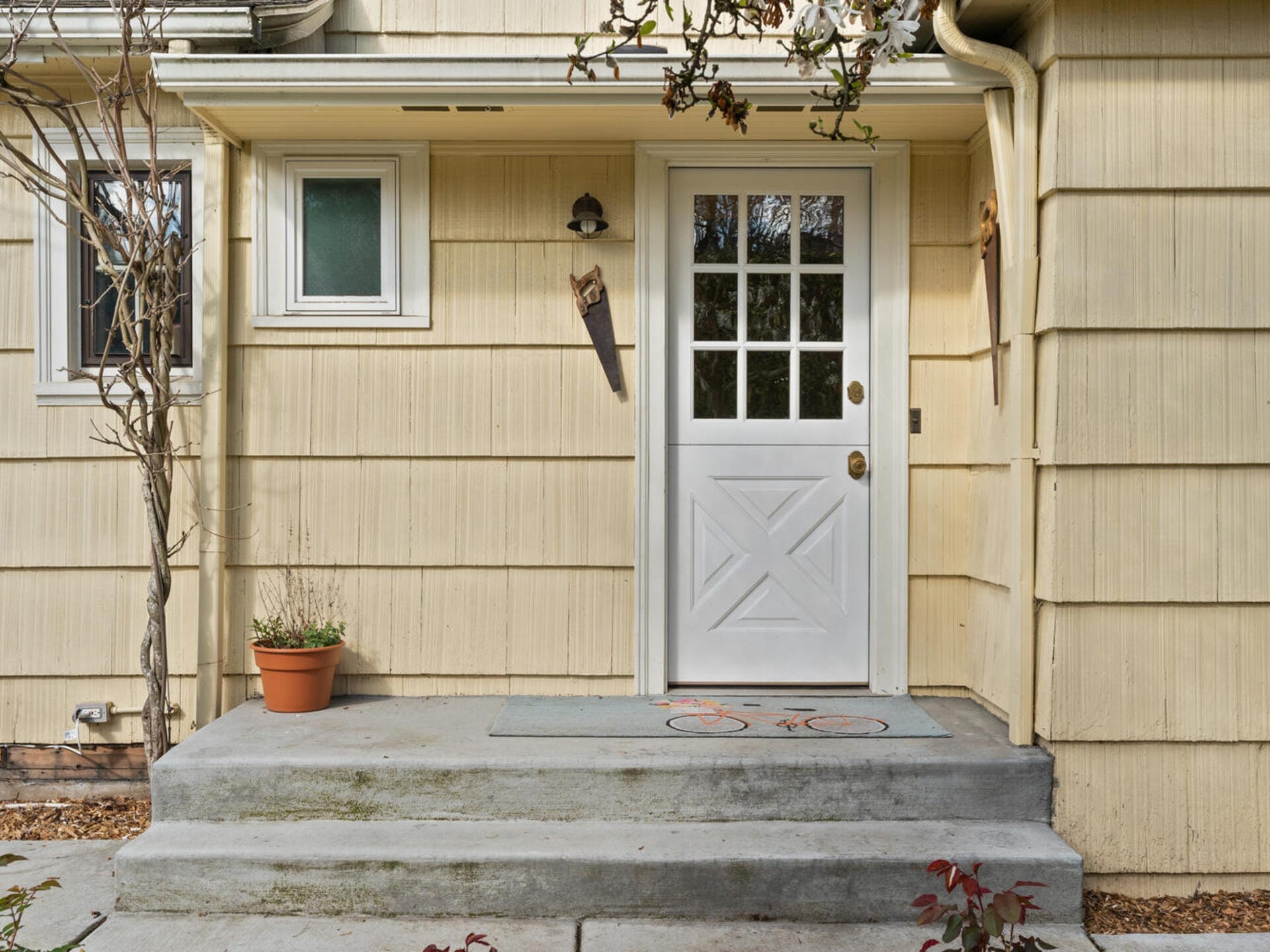 A yellow house with a white door featuring glass panels. The entrance has a small concrete porch with a potted plant and a doormat decorated with a bicycle design. Two decorative saws are mounted on the exterior walls.