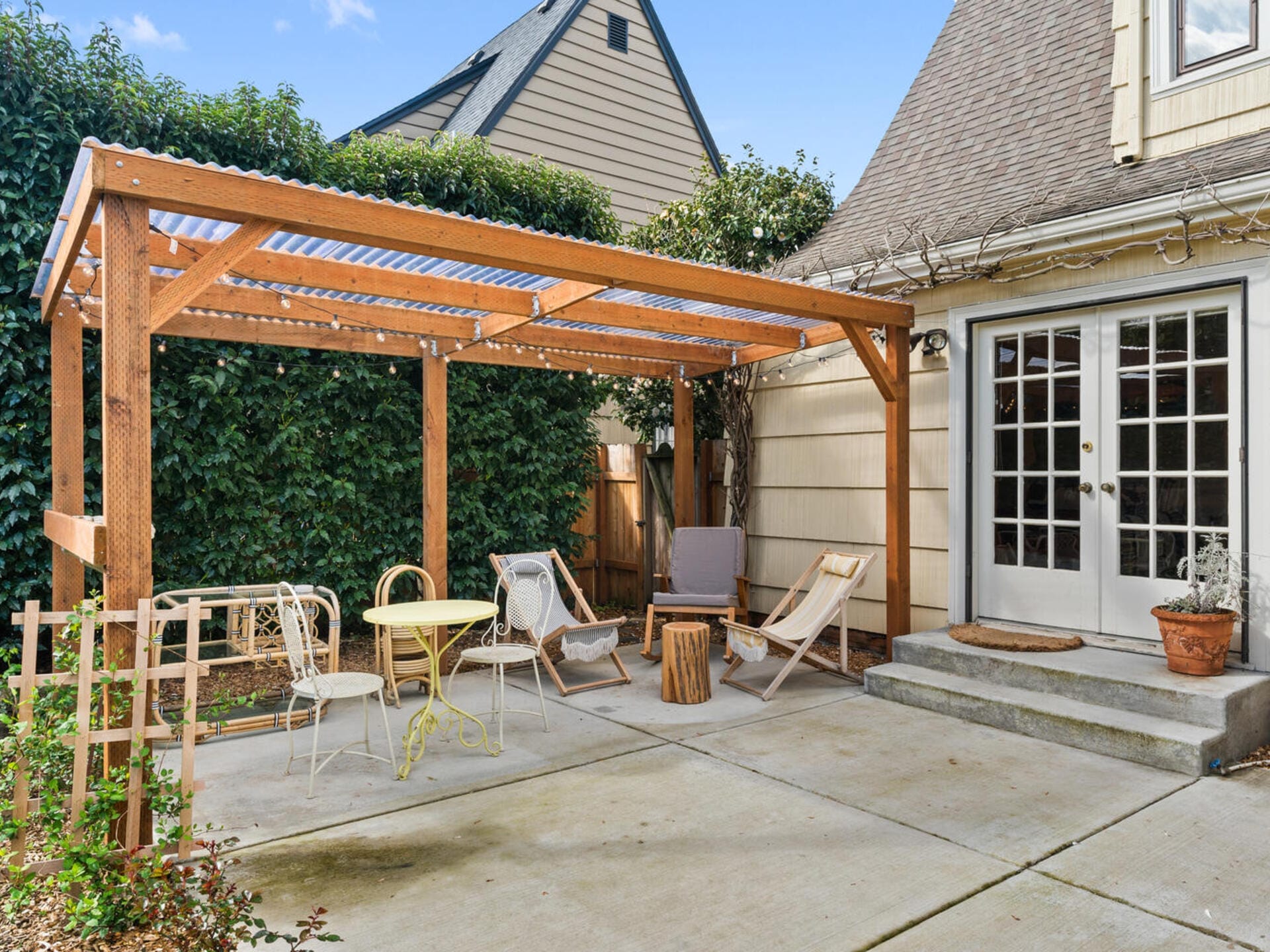A backyard with a wooden pergola over a concrete patio. The area features lounge chairs, a small yellow table, a wooden stump, and potted plants. French doors lead into the house, and a green hedge provides privacy.