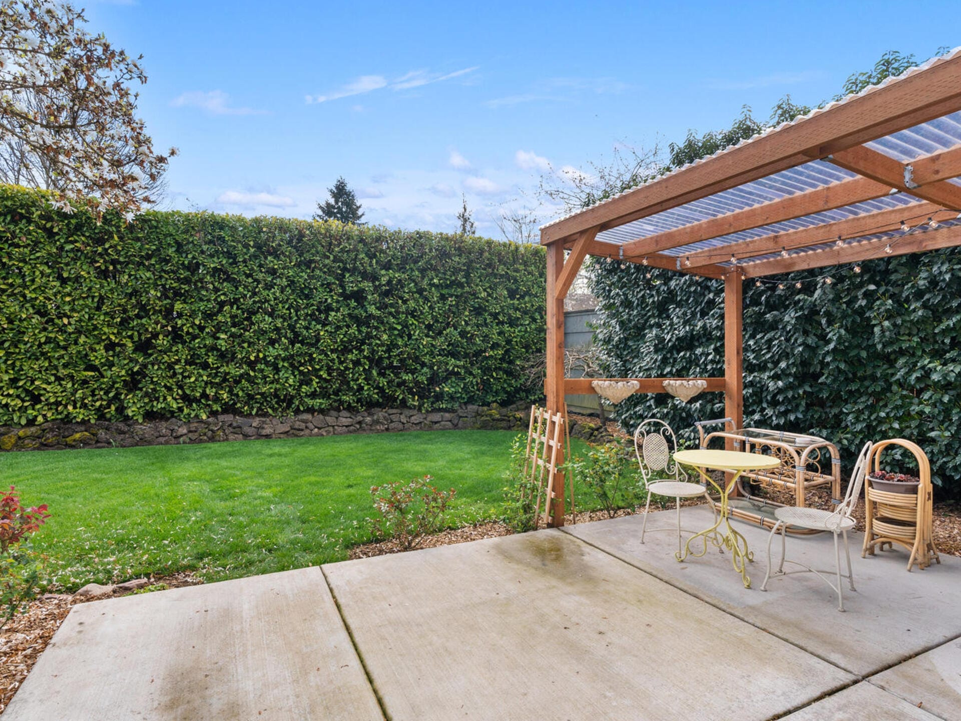 A backyard patio with a pergola covered in clear panels. It features a small round table and chairs. The patio overlooks a well-manicured lawn bordered by tall, dense hedges under a bright blue sky.