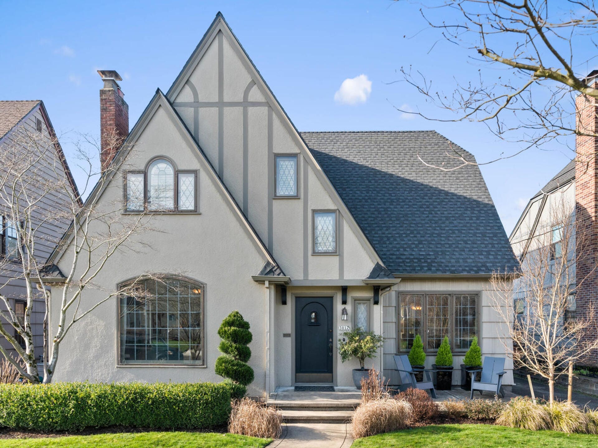 A charming Tudor-style home with a steep gabled roof, light gray stucco, and decorative trim. The front yard features manicured shrubs, a small tree, and a brick pathway leading to a dark gray front door with sidelights and a lantern.