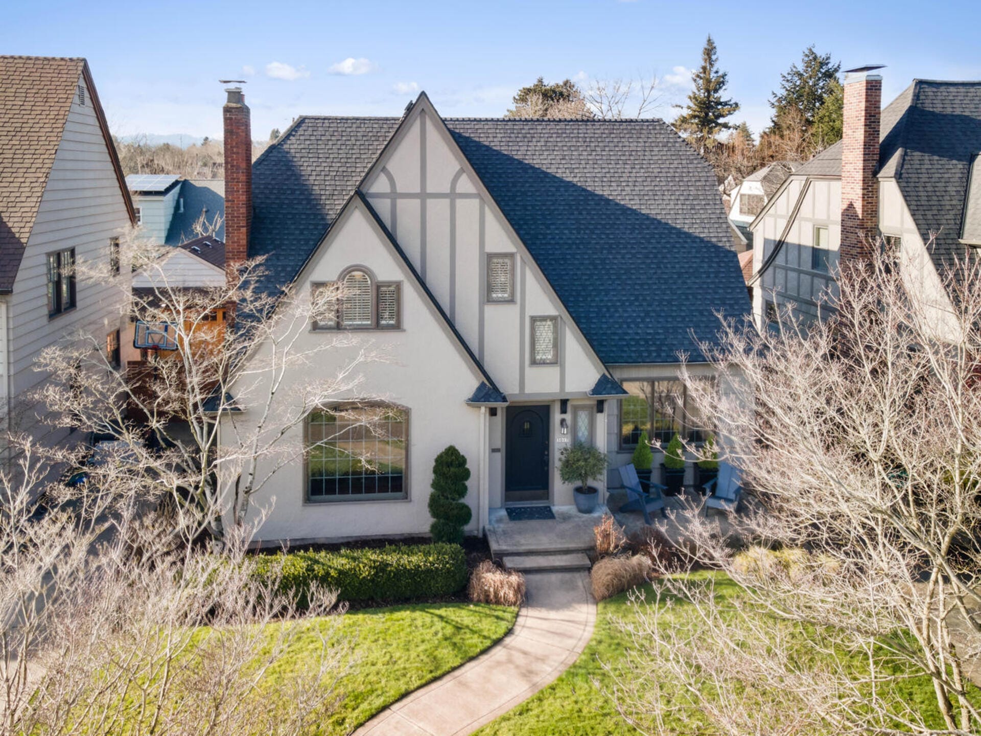 A charming two-story Tudor-style house with steeply pitched rooflines and white stucco exterior is surrounded by bare trees and a neatly manicured lawn. A curved walkway leads to the front entrance with a small porch and arched window above.