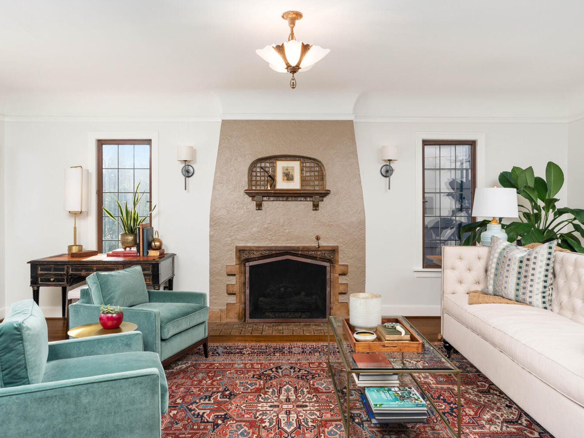 A cozy living room with two teal armchairs, a white sofa, and a decorative fireplace. There are modern lamps, a patterned rug, and a coffee table with magazines. Plants add a touch of greenery to the bright, inviting space.