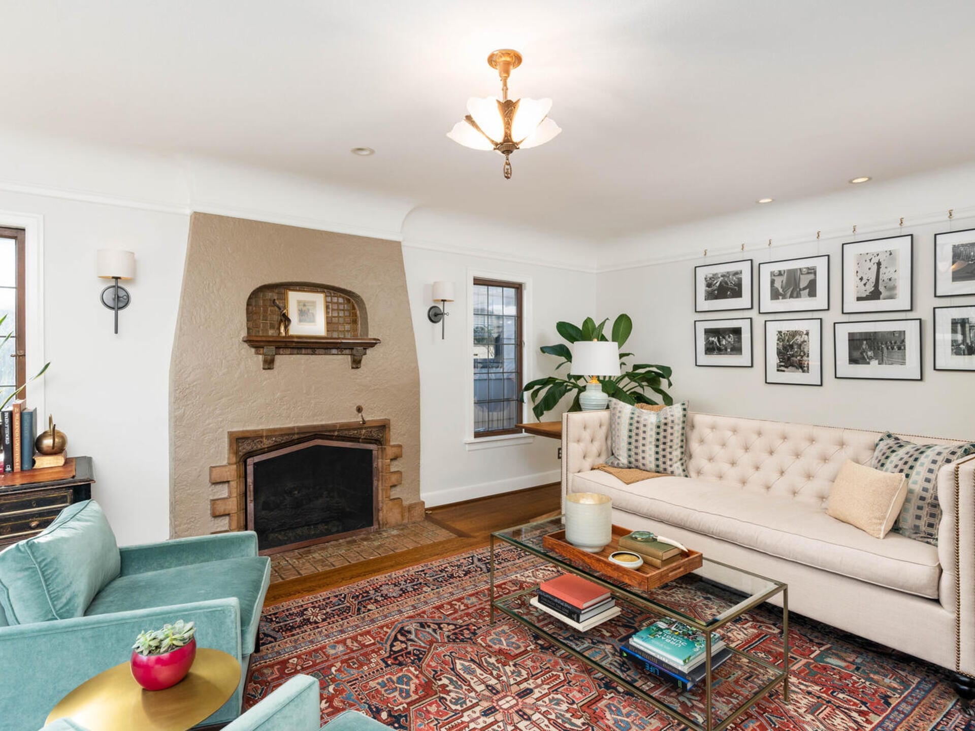 A cozy living room with a beige sofa, teal armchairs, and a glass coffee table over a patterned rug. The room features a rustic fireplace, various plants, and a wall decorated with black-and-white photographs. A chandelier hangs from the ceiling.