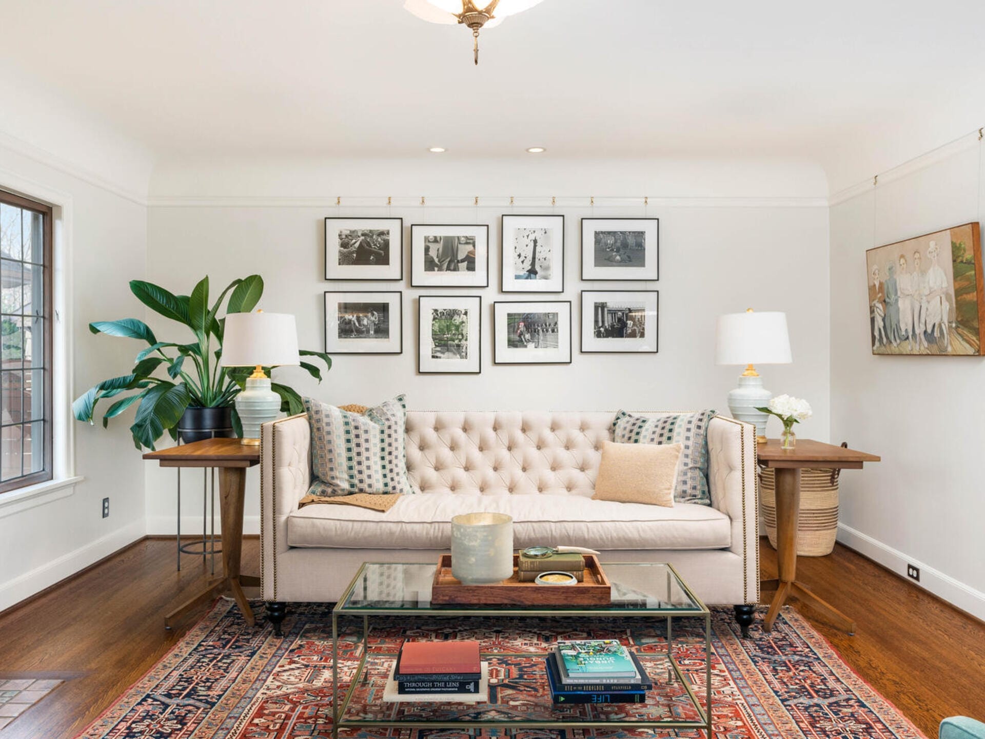 A cozy living room with a beige sofa, decorative pillows, and a glass coffee table on an ornate rug. Framed black-and-white photos hang above the sofa. A large potted plant is on the left and a basket with flowers on the right. Wood floors and ceiling light.