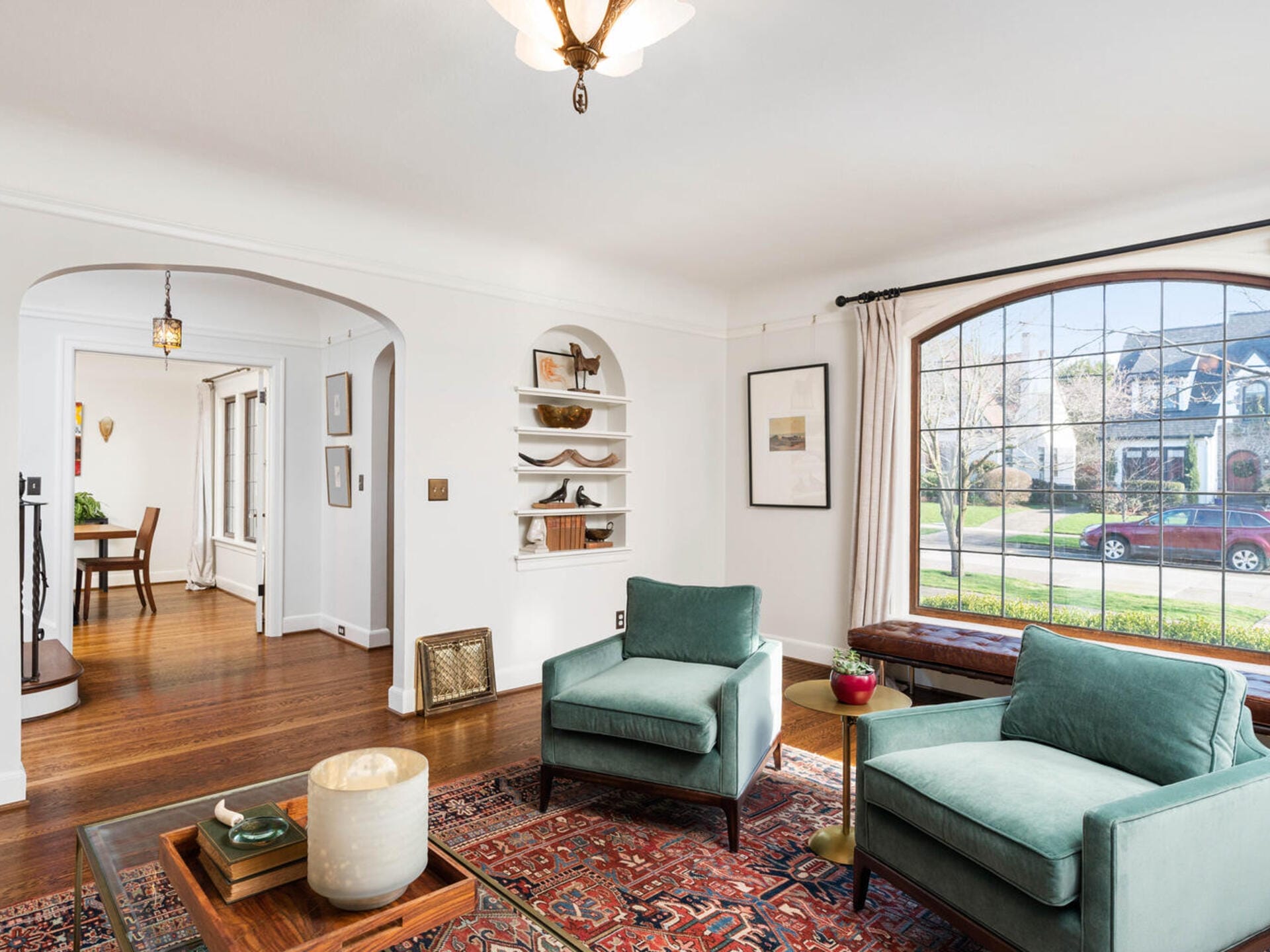 A cozy living room features two green armchairs on a patterned rug, a wooden coffee table, and a large arched window offering a street view. Shelves with decor and wall art add charm. An open doorway leads to a dining area with hardwood floors.