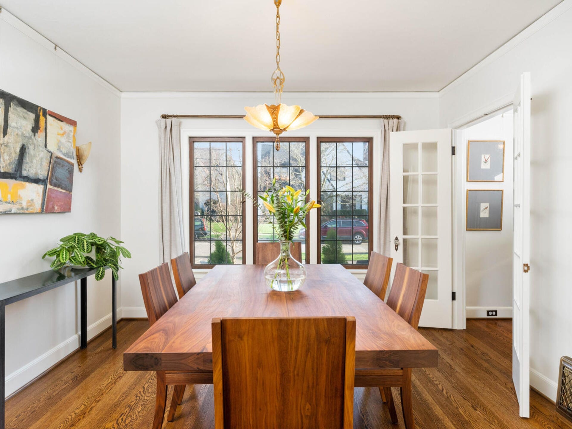 A bright dining room with a wooden table set for six, a vase of yellow flowers as the centerpiece. Large windows with sheer curtains let in natural light. A modern painting and a potted plant decorate the space.