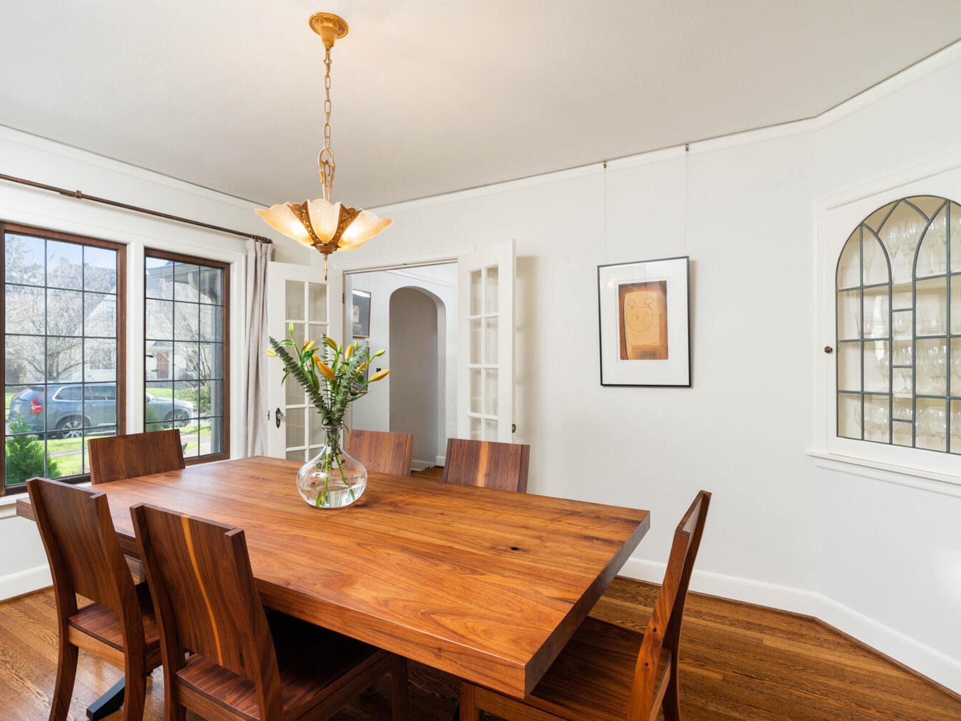 A dining room with a wooden table and chairs, a glass vase with flowers on top. There are glass double doors, a large window with a view of greenery, a hanging light fixture, and wall art. The floor is hardwood.