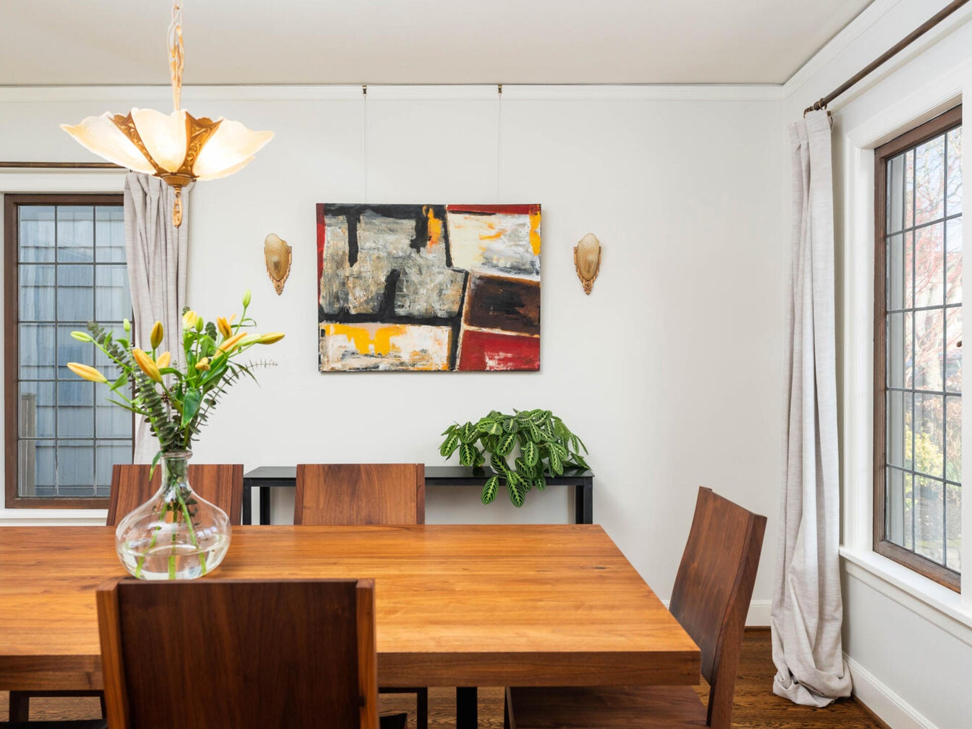 A dining room with a wooden table and chairs is warmly lit by a hanging light fixture. A vase with flowers sits on the table. An abstract painting and a potted plant on a black shelf adorn the white wall. Large windows with curtains are on the sides.