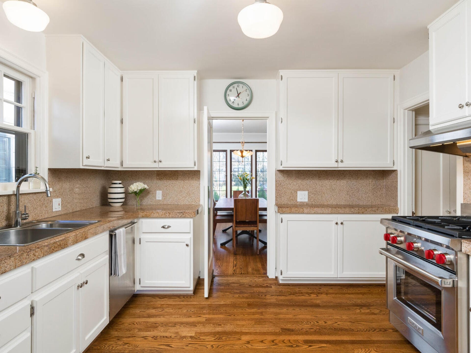 A modern kitchen with white cabinets, granite countertops, and stainless steel appliances. A clock is on the wall above the doorway leading to a dining room with a wooden table and chairs. The floor is hardwood. Bright and tidy atmosphere.