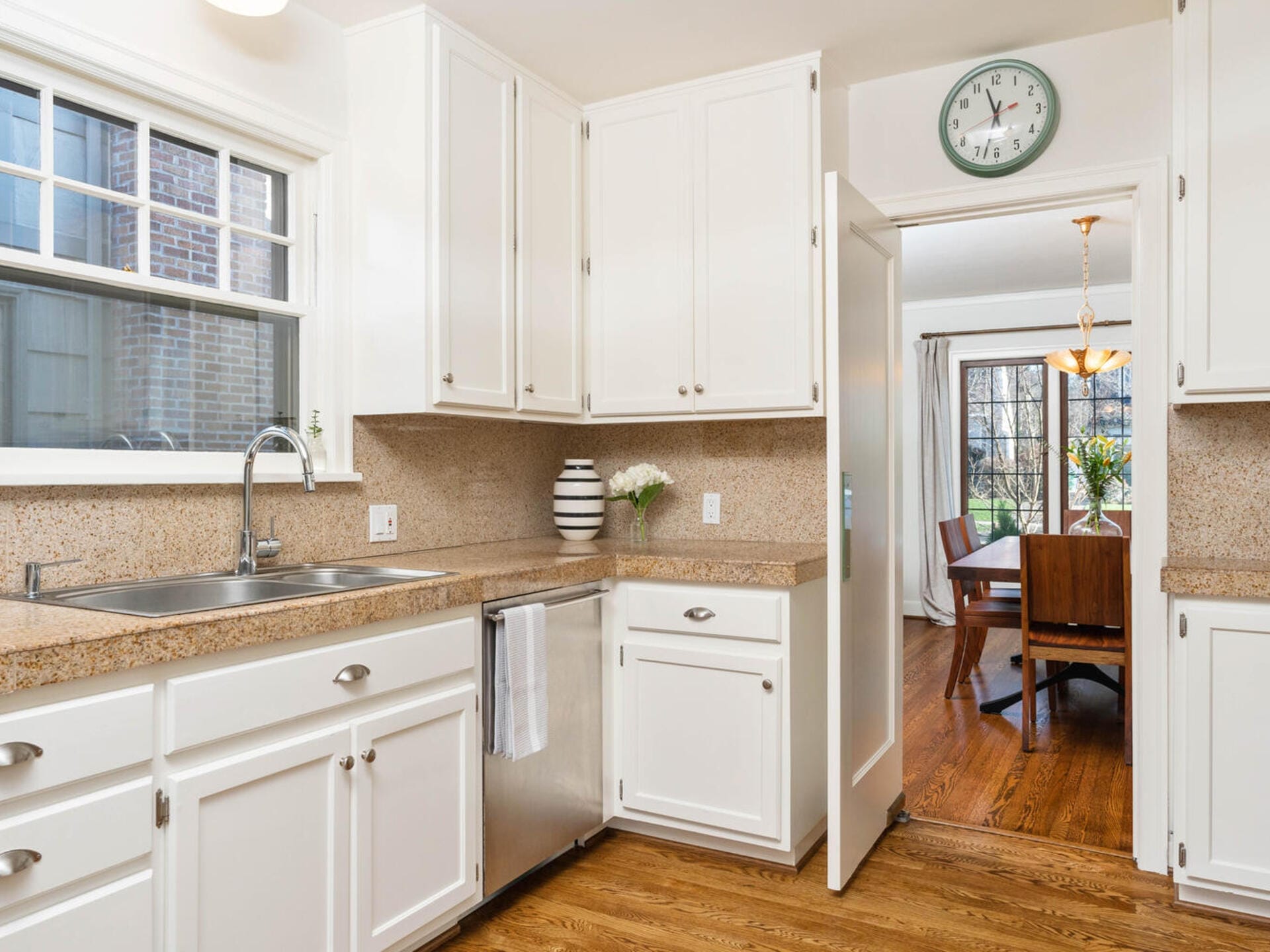 A bright kitchen with white cabinets and a light brown countertop. Theres a stainless steel sink, a towel hanging, and a vase with flowers. A clock hangs above a doorway leading to a dining area with a wooden table and chairs, and a window view.