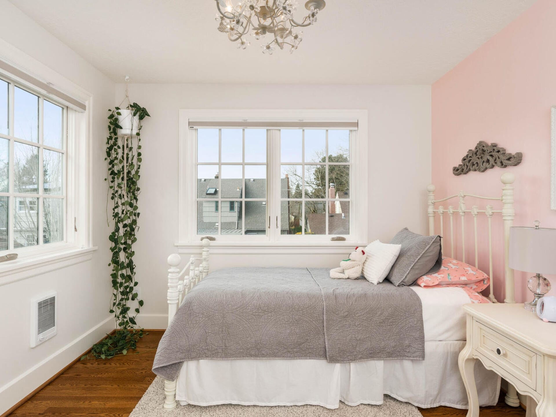 A cozy bedroom with a white metal bed frame, gray and pink bedding, and a pink accent wall. A plant hangs near two large windows, bringing in natural light. A chandelier hangs from the ceiling, and a small nightstand with a lamp sits beside the bed.