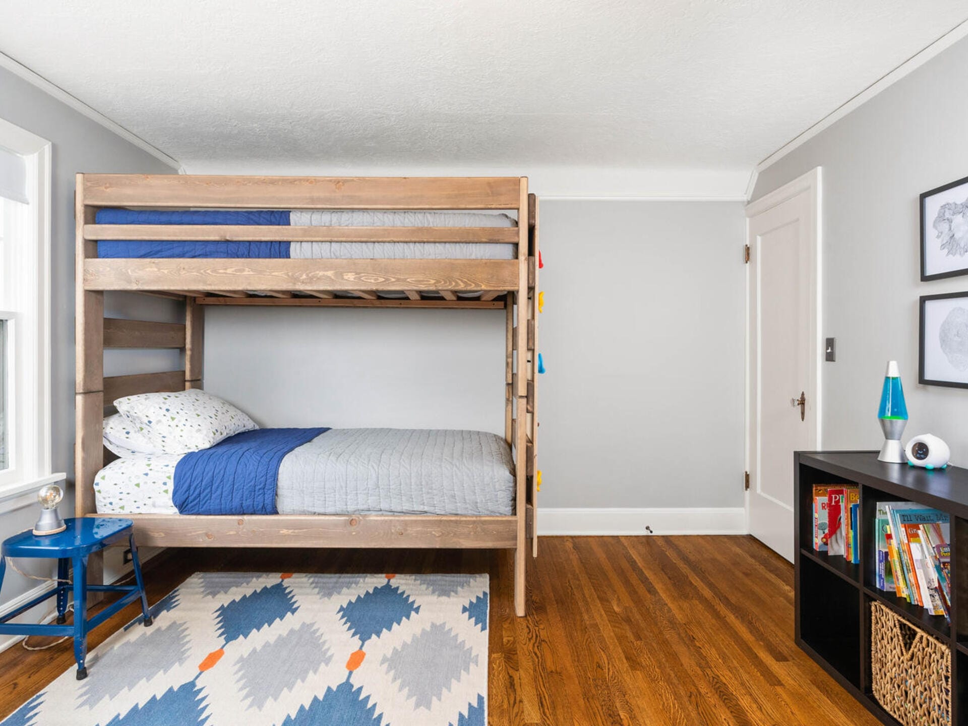 A neatly organized bedroom features wooden bunk beds with blue and gray bedding. A blue side table is beside the lower bunk. The room has a colorful geometric rug and a black shelf with books and decor. Walls are light gray with framed artwork.