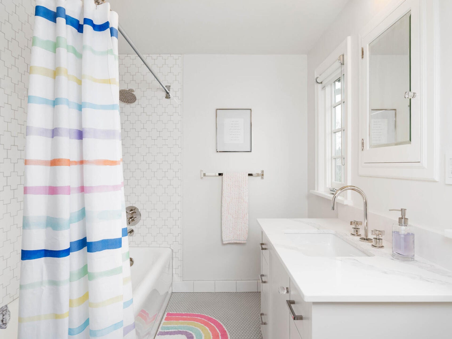 A bright bathroom featuring a white bathtub with a colorful striped shower curtain, a white vanity with a sink and silver faucet, a mirror above, a framed picture on the wall, and a striped bath mat on the floor.