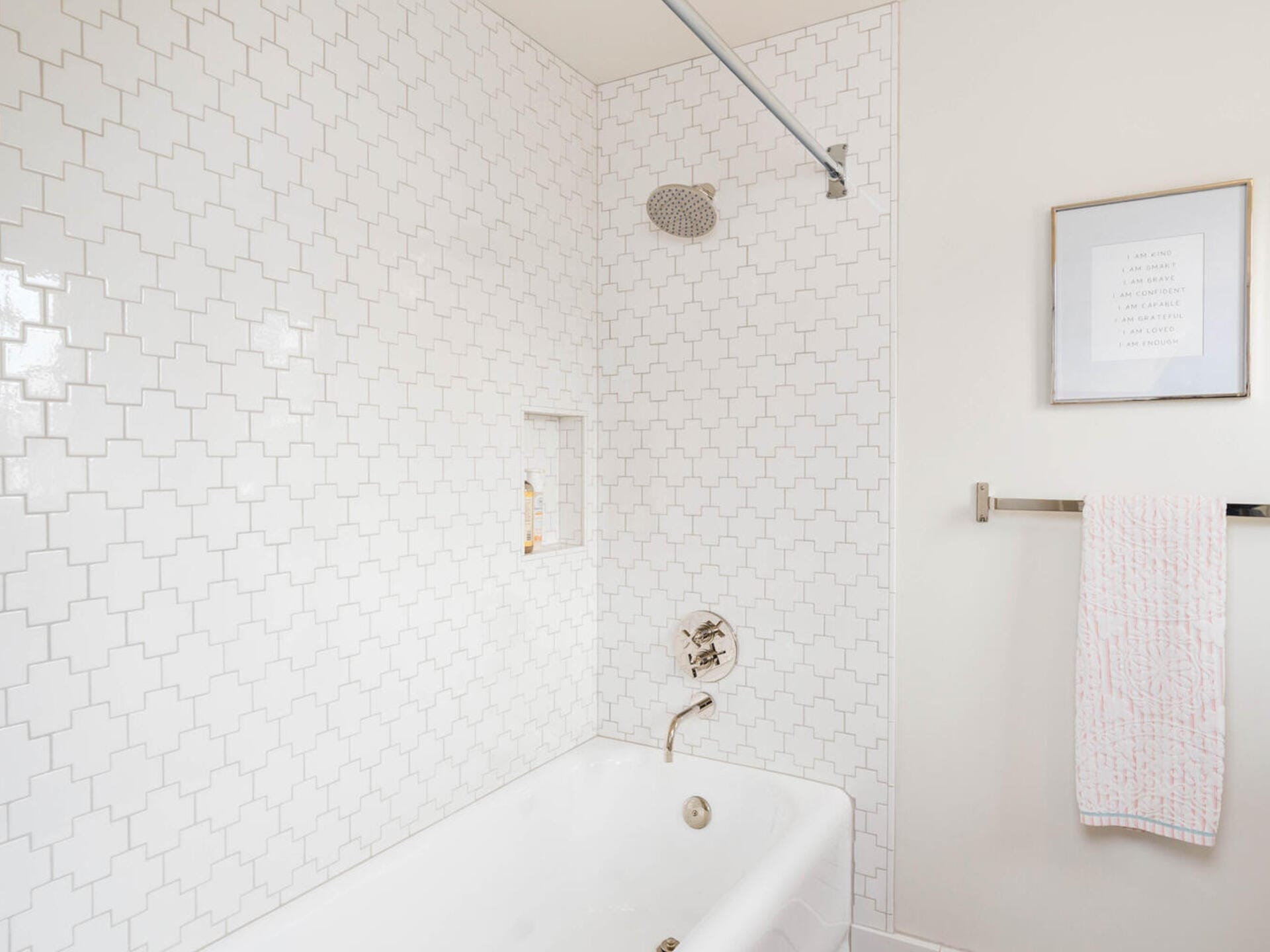 A minimalist bathroom with a white bathtub and geometric patterned tiles on the wall. A silver showerhead is above the tub, and a niche holds toiletries. A towel hangs on a nearby rack, and a framed picture is on the wall.