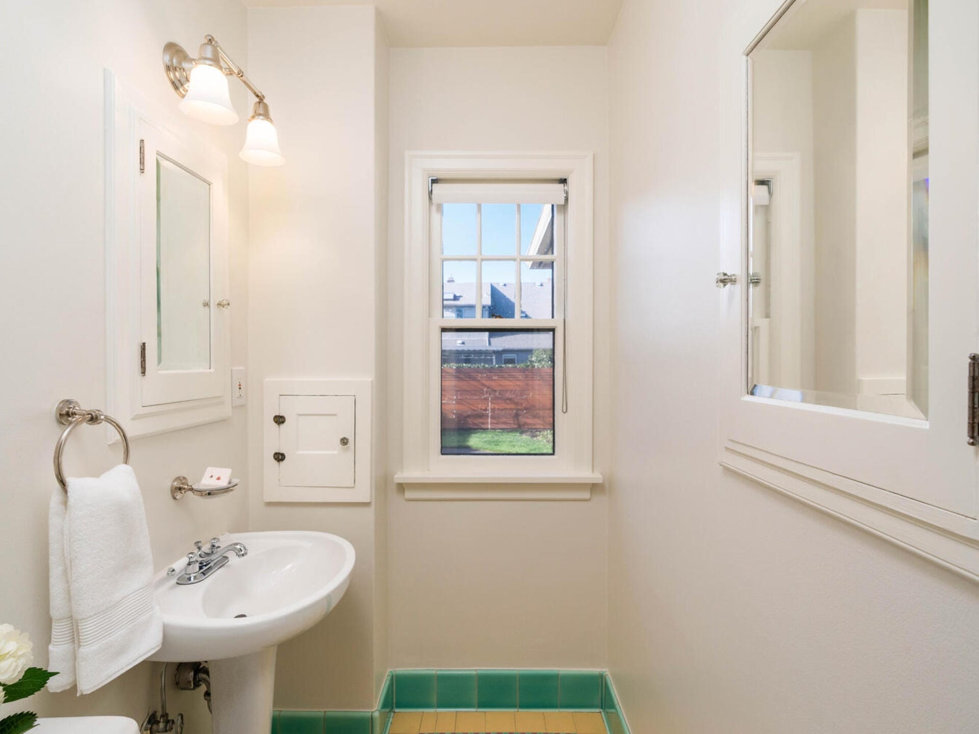 A small bathroom with a white sink and towel holder on the left wall. Above the sink are two wall lights. A window with white trim is centered on the far wall. The room has white walls and a beige-tiled floor with a green border.