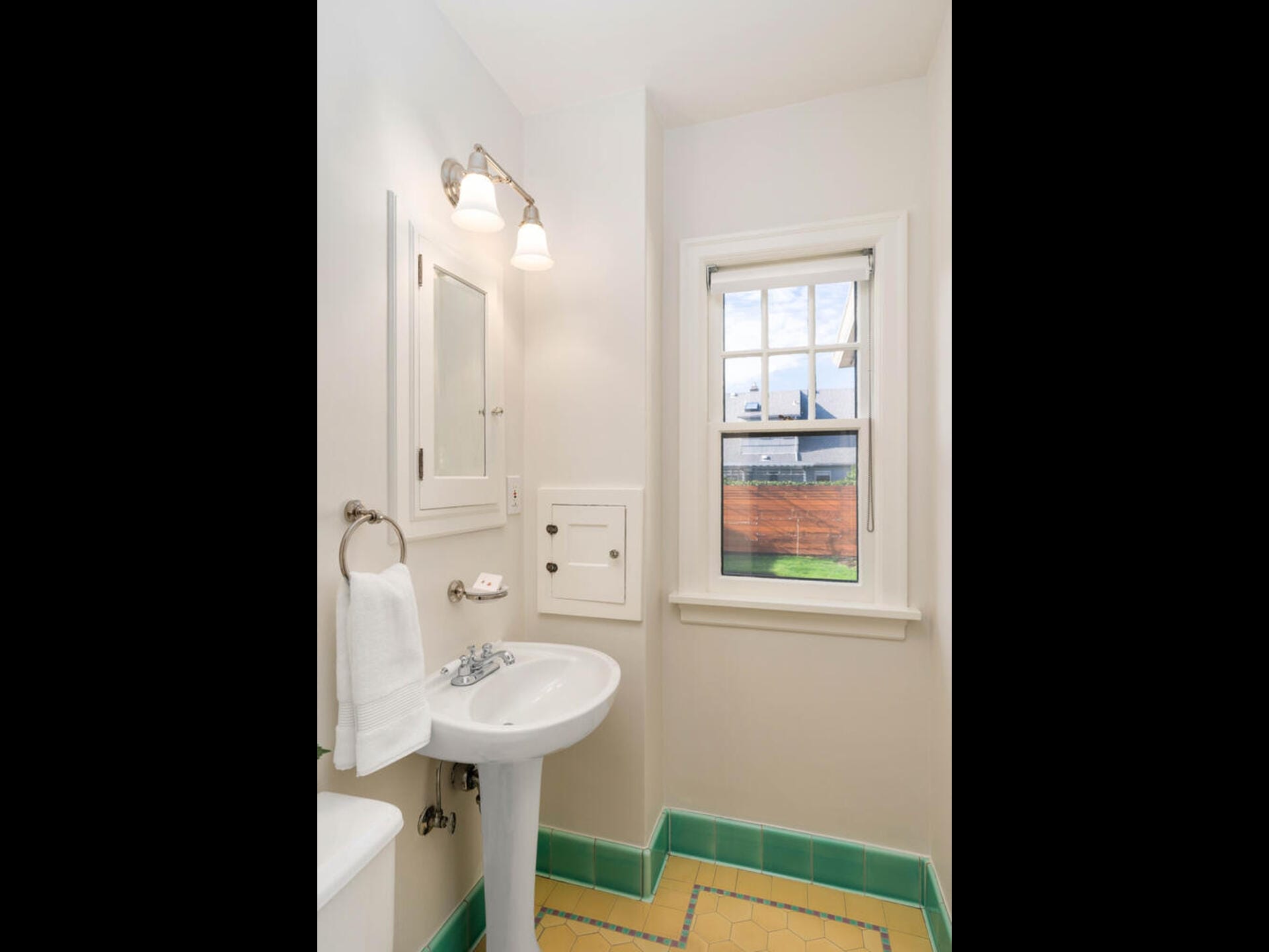 A bright bathroom with a pedestal sink, towel ring with a white towel, wall-mounted mirror cabinet, and double light fixture. The floor features yellow and green tiles, and a window shows an outdoor view with a wooden fence and greenery.