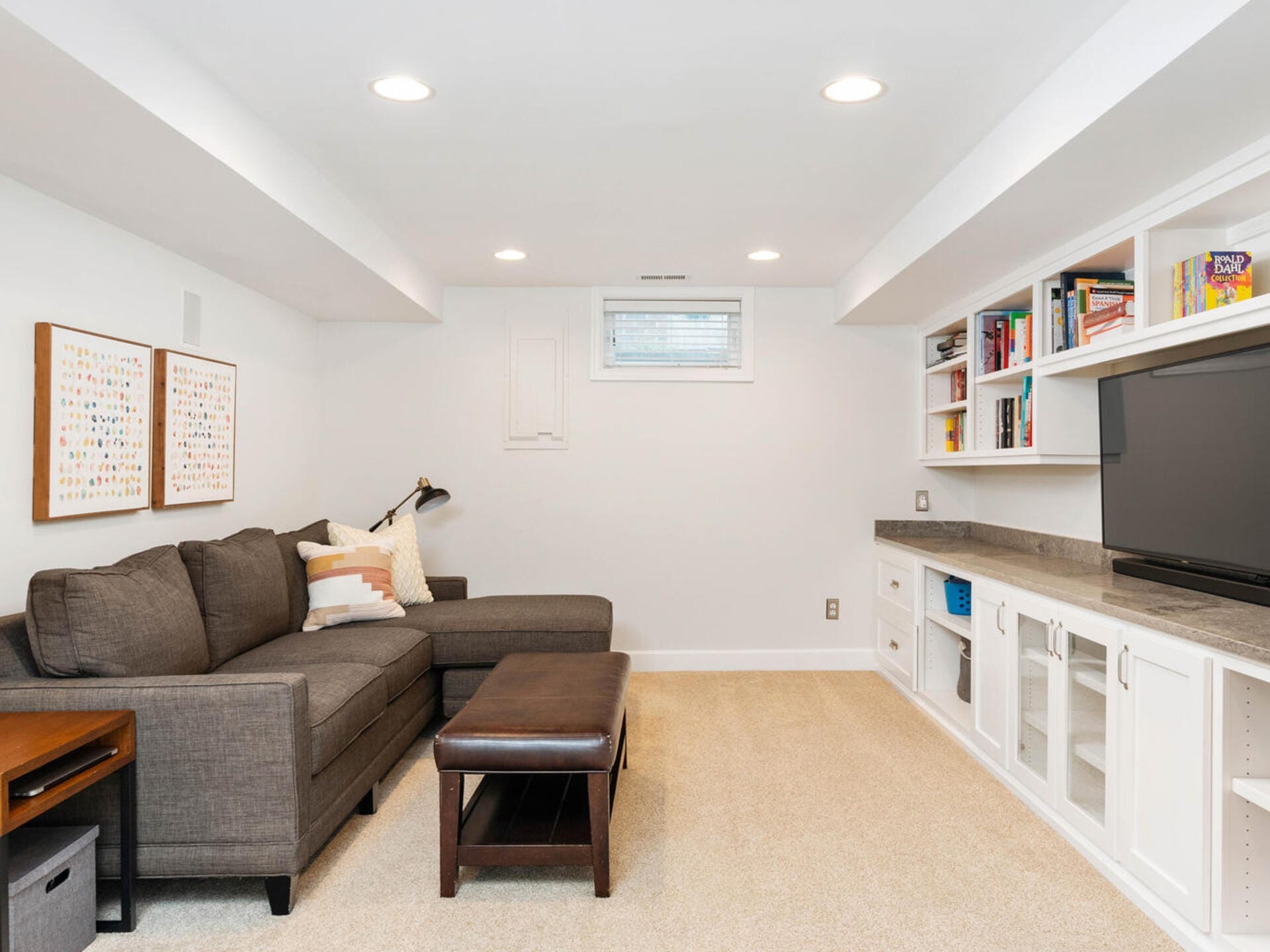 A cozy basement living room with a gray sectional sofa, brown ottoman, and a floor lamp. Built-in shelves and cabinets hold books and games. A large TV is mounted on the wall. The room is carpeted with neutral tones and has recessed lighting.