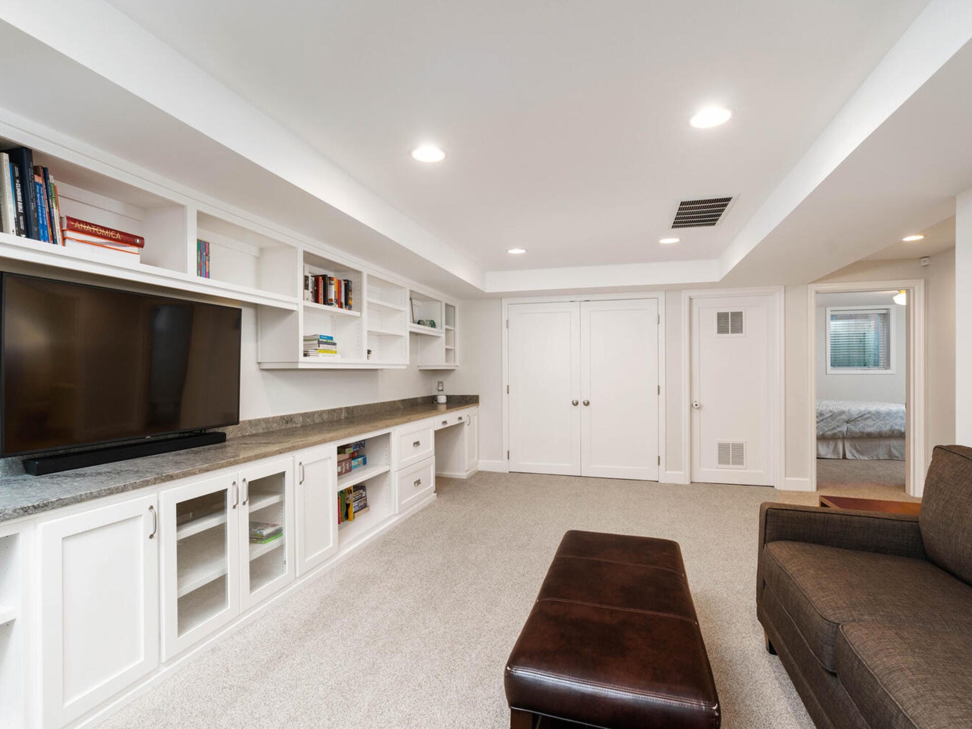 Spacious basement room with white built-in shelves and cabinets, a large flat-screen TV, and a brown leather ottoman. The room has carpeted flooring and recessed lighting, with a doorway leading to another room with a bed.