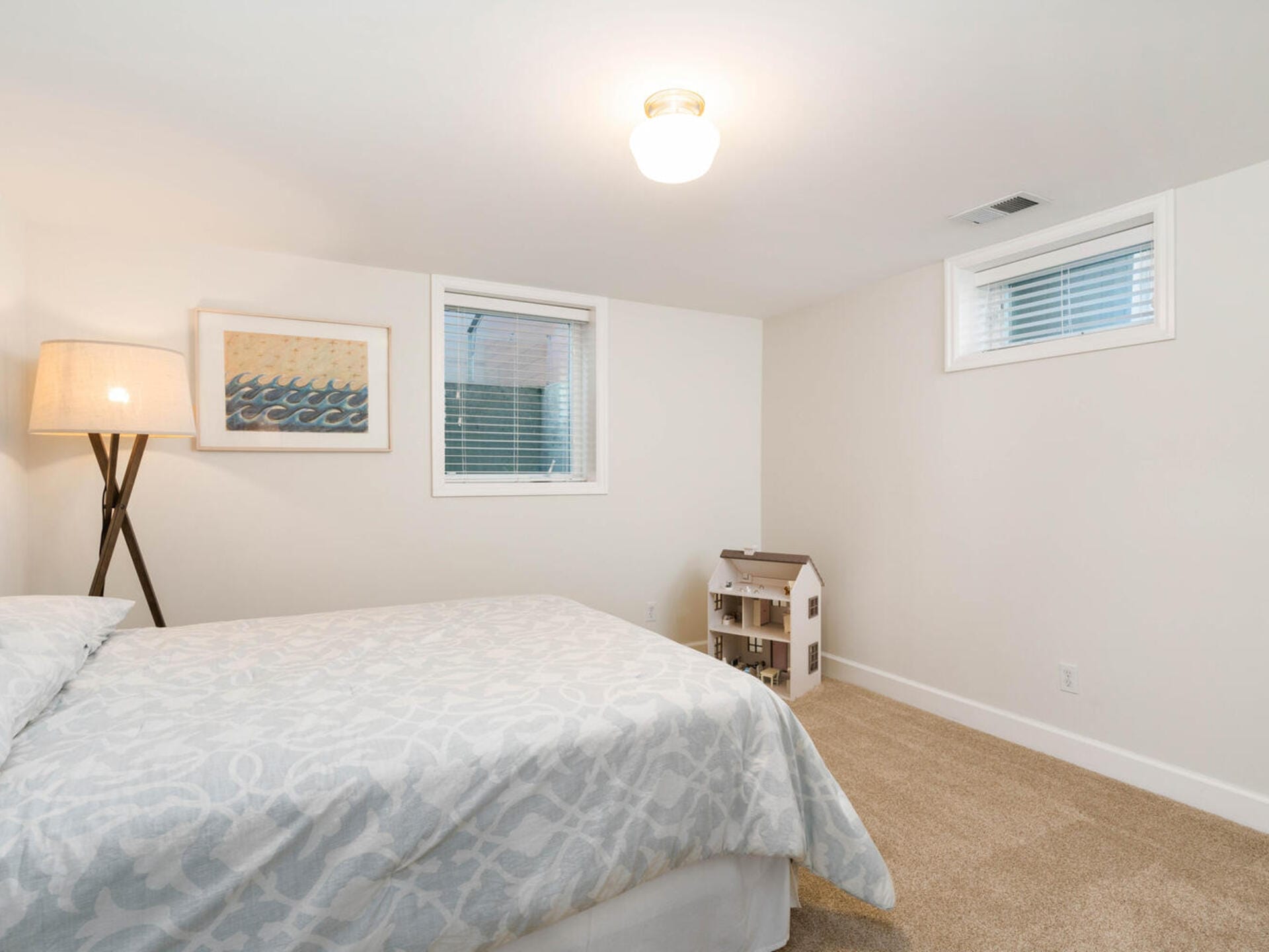 A minimalistic bedroom features a bed with a patterned duvet, a floor lamp, a framed print on the wall, and a small dollhouse in a corner. The room has carpeted flooring, two small windows, and soft lighting.