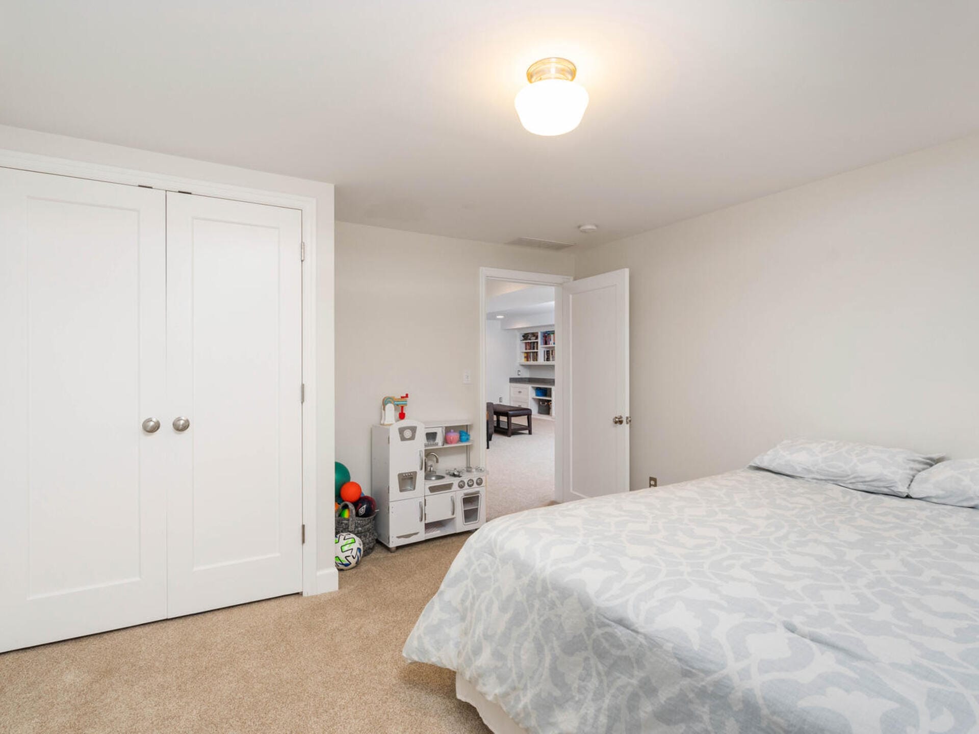 A bedroom with a large bed covered in a patterned duvet, a double-door closet, and a door leading to another room. The carpet is beige, and theres a small toy kitchen set with colorful balls in the corner. Walls are painted white.