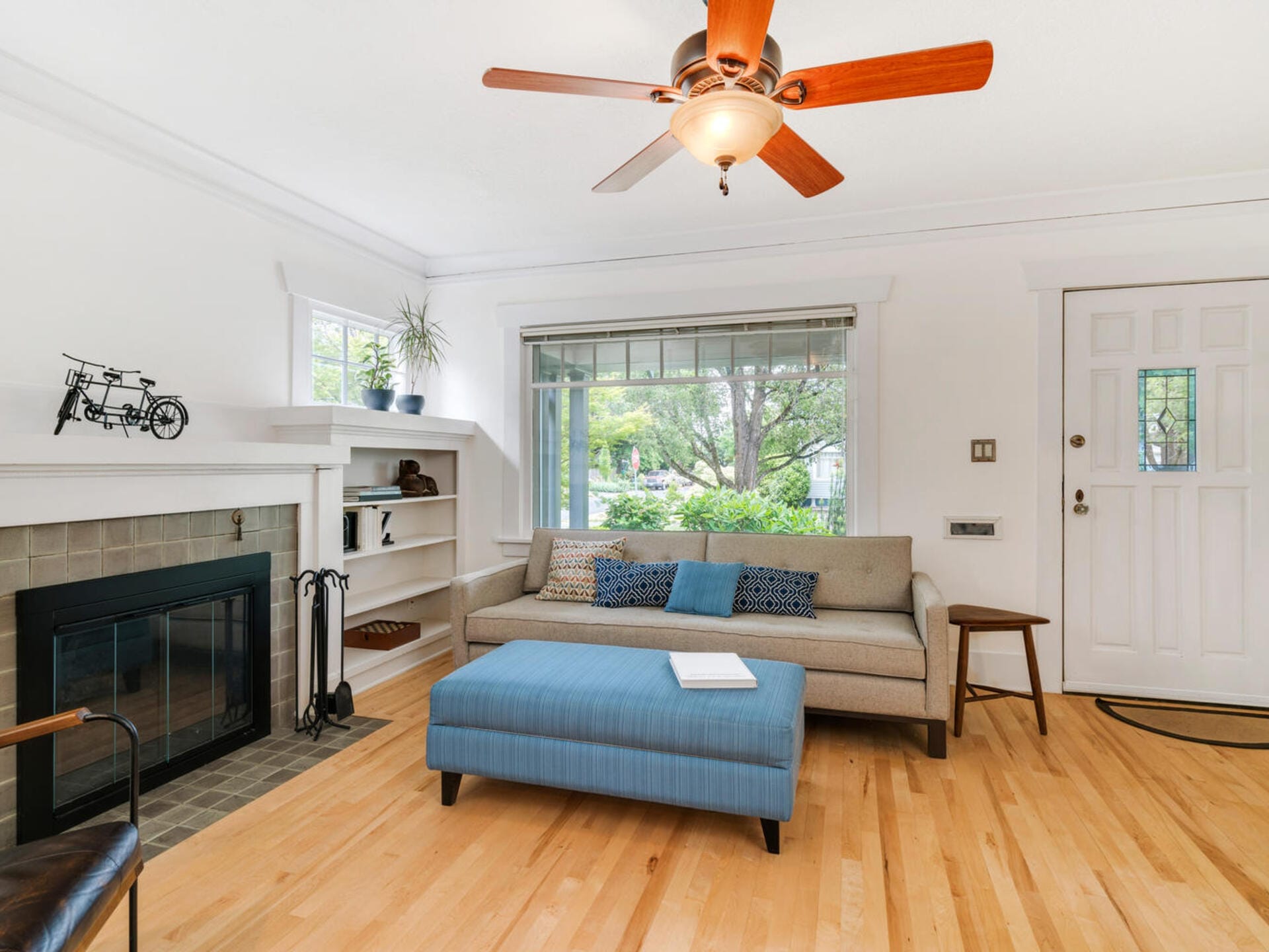 A cozy living room with a beige sofa, a blue ottoman, and a fireplace. A ceiling fan with wooden blades hangs above. Shelves with decor items and a plant sit next to a sunlit window with a view of greenery outside. Hardwood floors complete the scene.