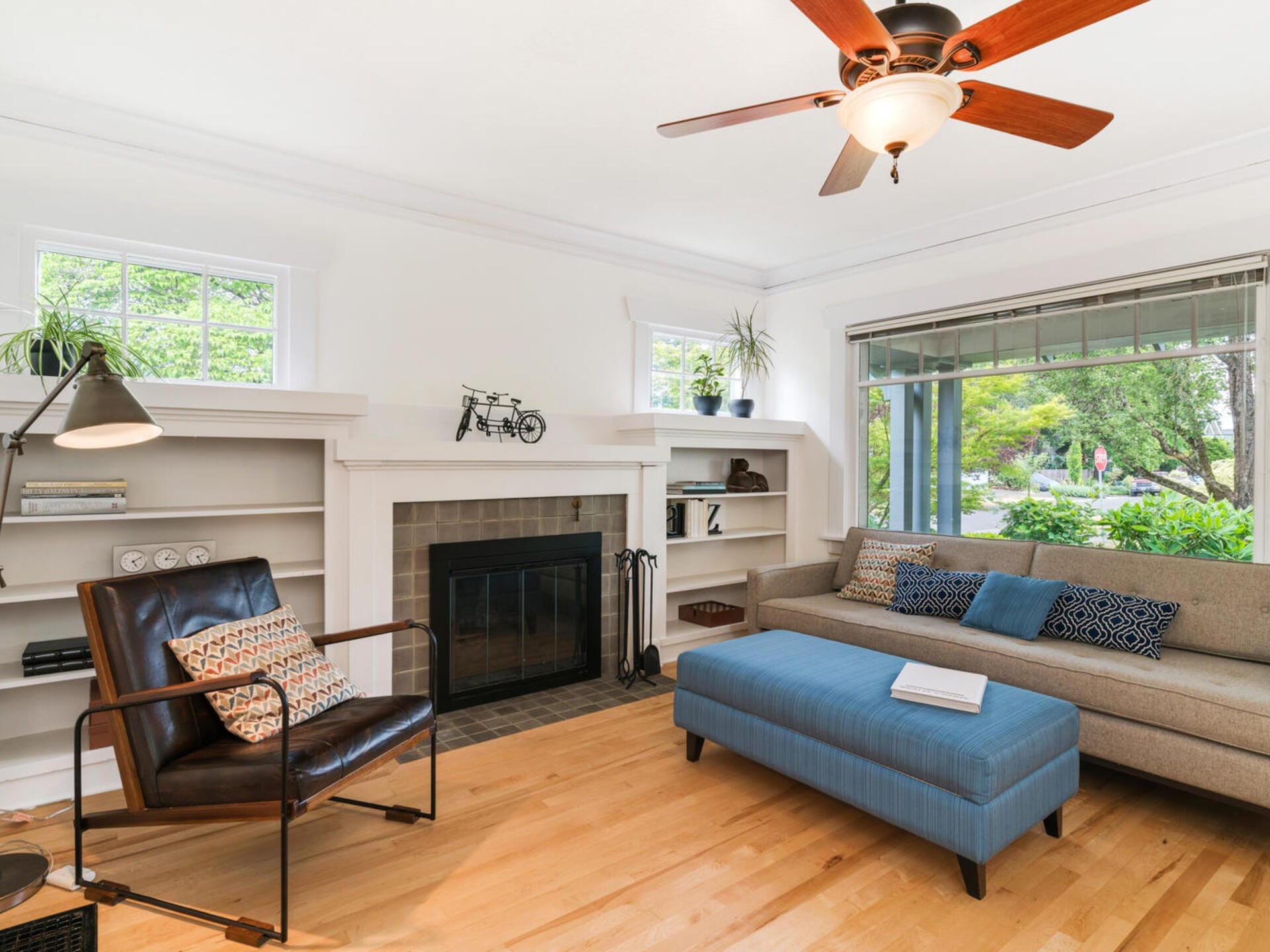 A cozy living room with a leather armchair, a gray couch with cushions, and a blue ottoman. There’s a fireplace with built-in shelves and a lamp nearby. Large windows offer a view of green foliage outside. A ceiling fan is overhead.