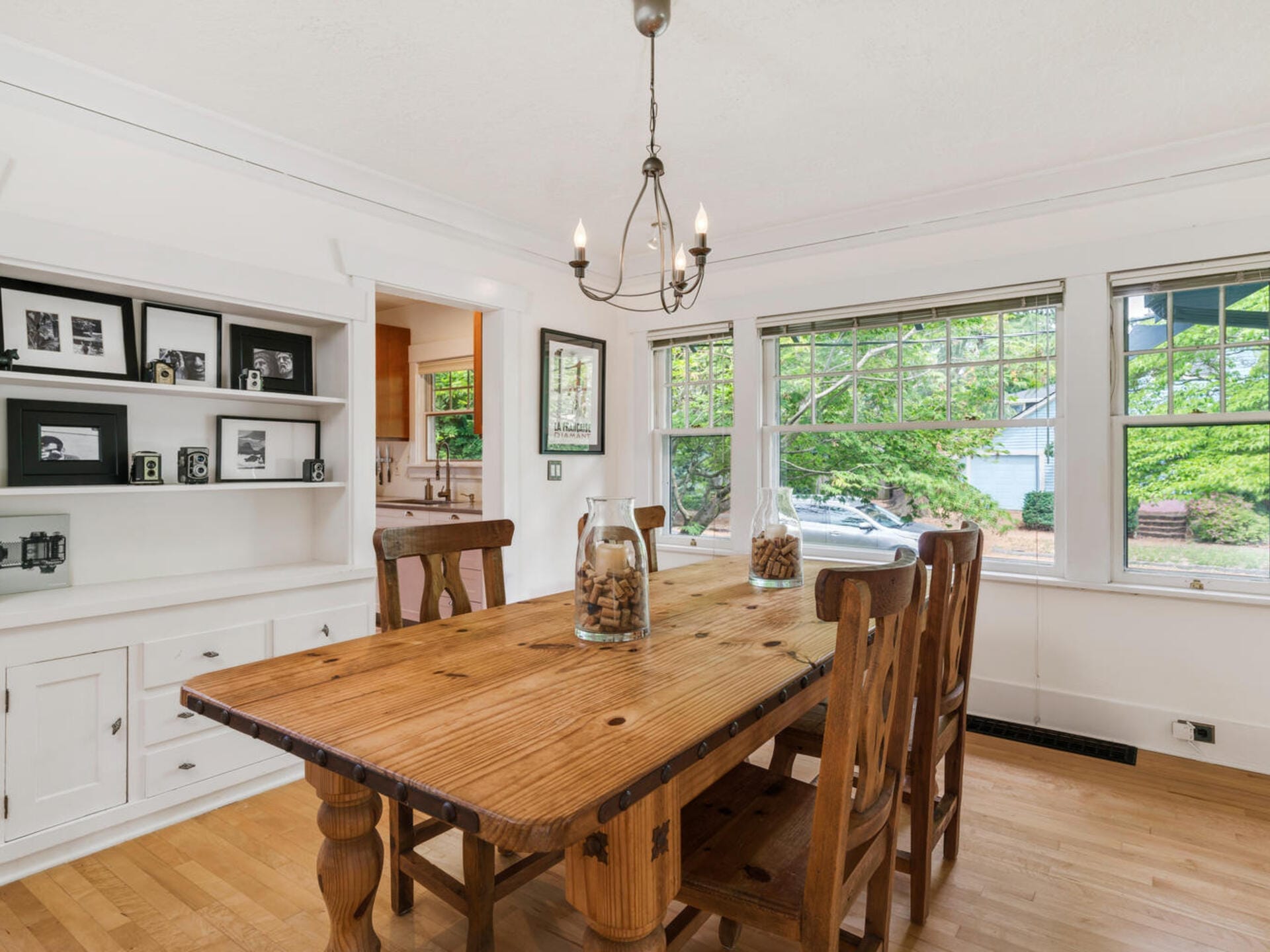 A bright dining room with a wooden table and six chairs. A chandelier hangs above the table, which is decorated with jars. Large windows offer a view of greenery outside. Built-in shelves display framed photos, and theres an adjacent door to the kitchen.