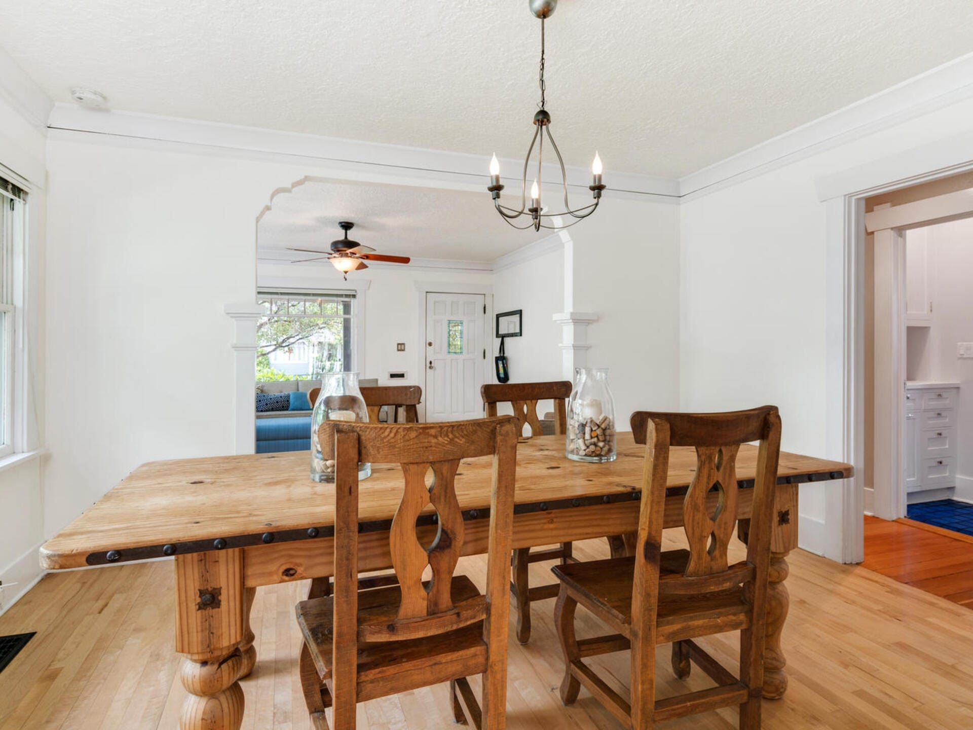 A dining room with a rustic wooden table and three matching chairs on a light wood floor. A chandelier hangs above the table. In the background, a doorway leads to a living room with a ceiling fan and a blue couch. Walls are painted white.