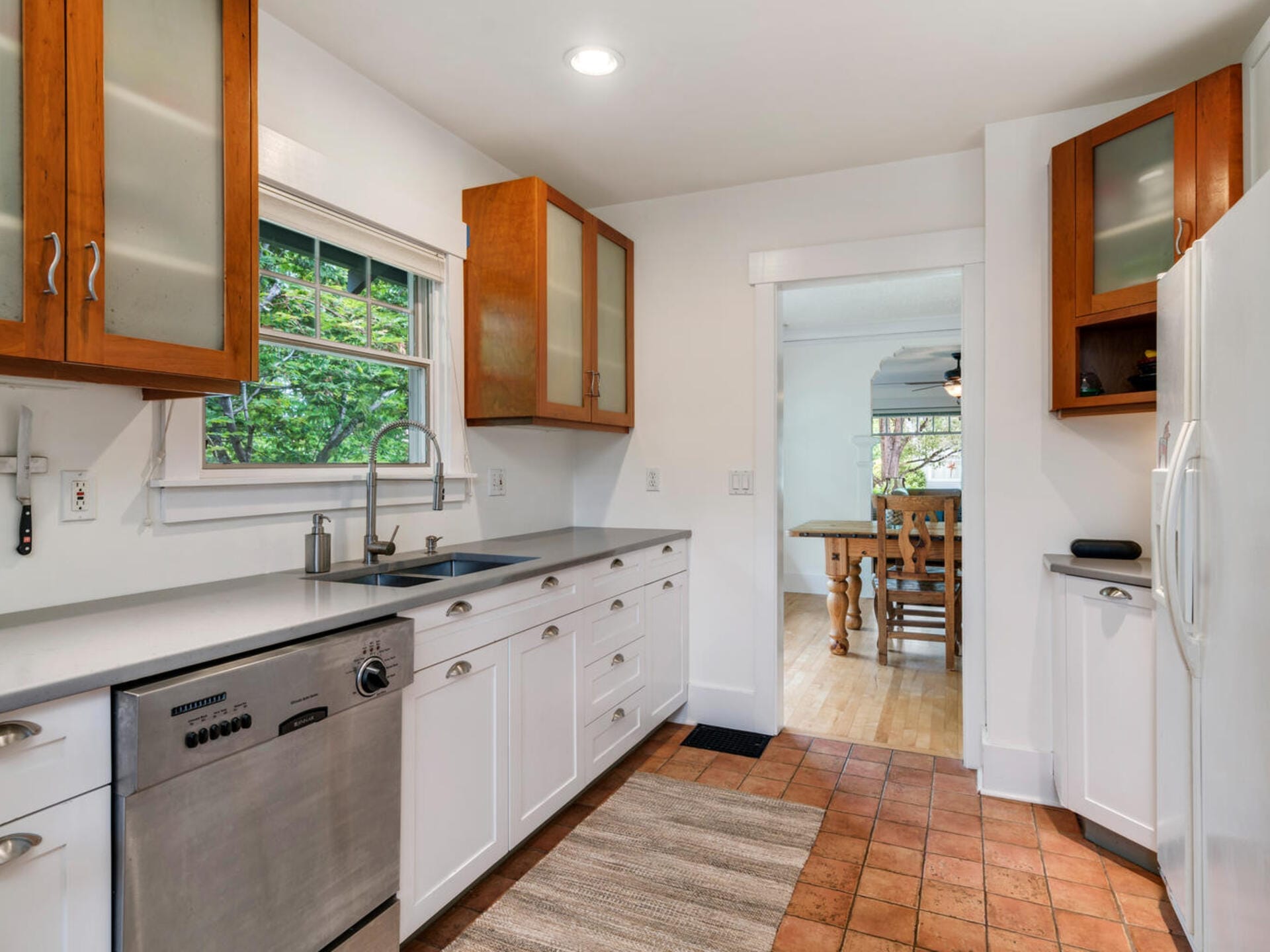 A modern kitchen with white cabinets and stainless steel appliances, including a dishwasher and refrigerator. Wooden upper cabinets and a window overlook greenery. Theres a striped rug on terracotta tiled flooring. A dining area is visible in the background.