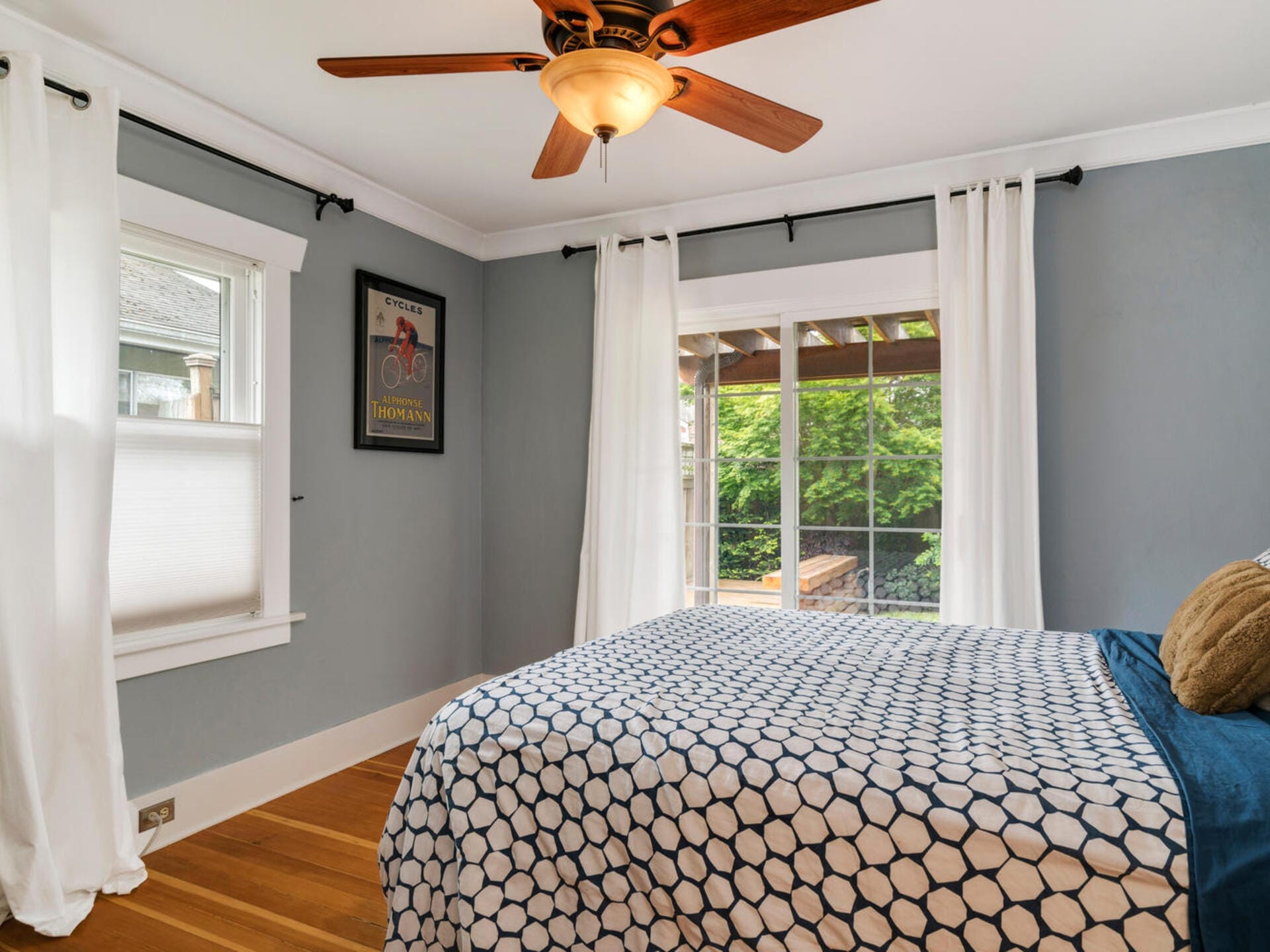 A cozy bedroom with blue walls and a wooden floor. A bed with a blue and white geometric-patterned comforter is beside large windows with sheer white curtains. A ceiling fan hangs above, and a framed poster is on the wall.