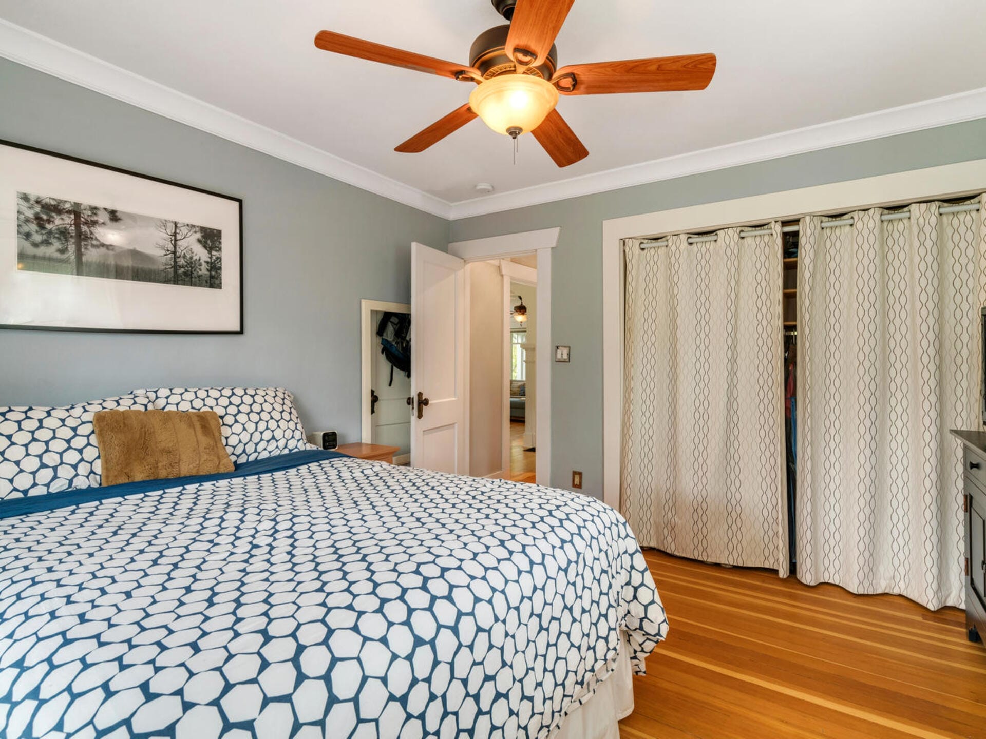 A cozy bedroom with a bed featuring blue and white geometric bedding. A ceiling fan with wooden blades is above. A framed landscape photo is on the wall, and a closet with curtains is on the side. The floor is wood, and the door is slightly open.