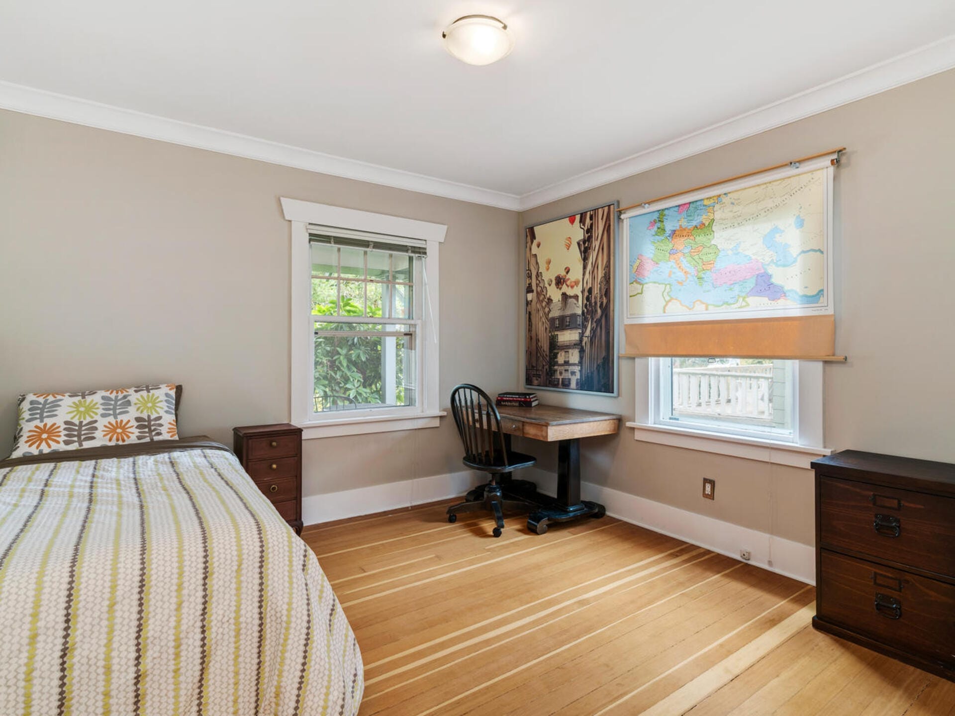 A cozy bedroom with a simple twin bed, a wooden desk with a black chair, and a window with a world map curtain. The room has a hardwood floor and neutral-colored walls, with a small dresser beside the bed. Natural light fills the space.