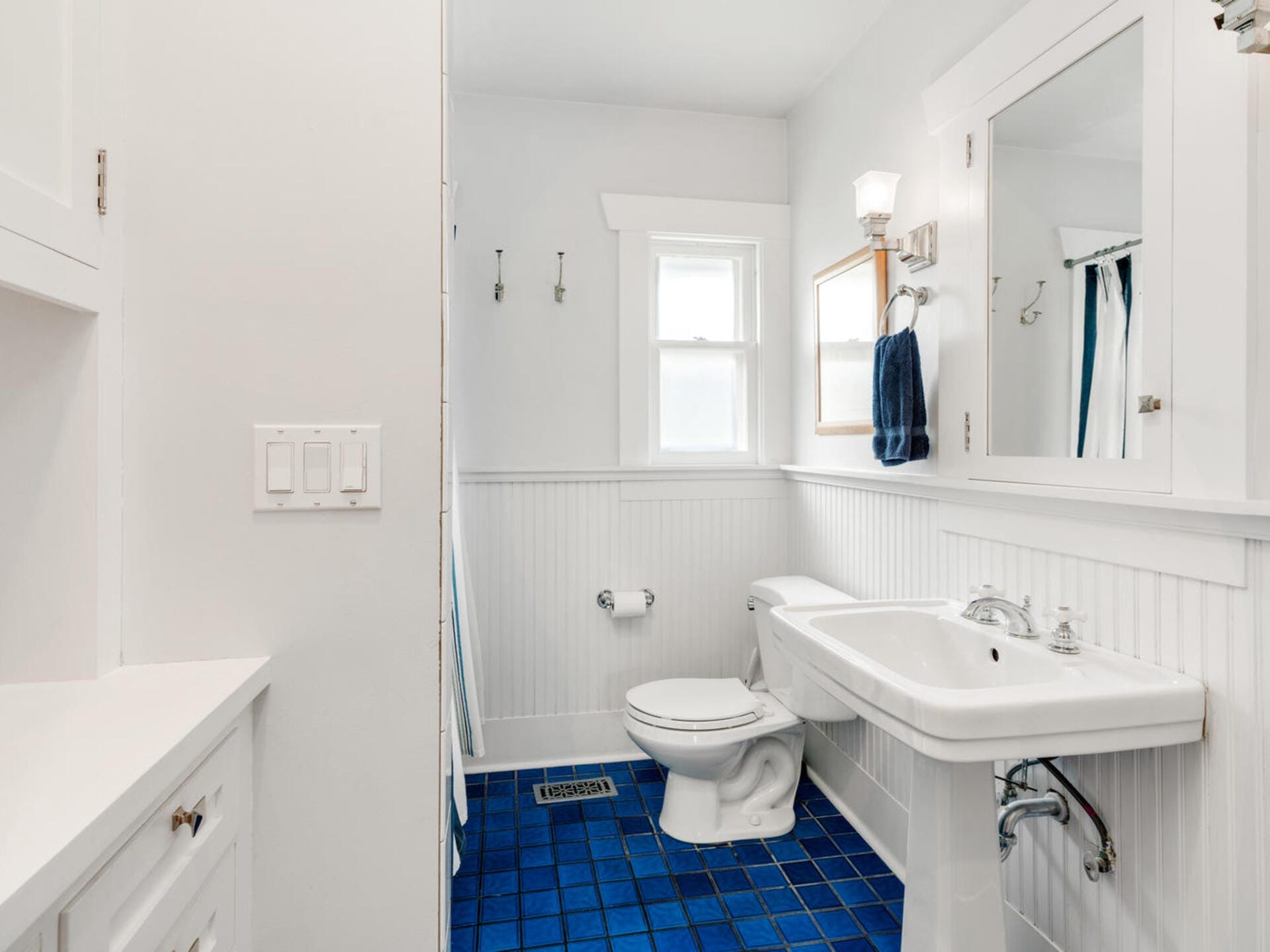 A bright bathroom with white walls and wainscoting, featuring a white pedestal sink and toilet, blue tiled floor, and a blue towel on a rack. A mirrored cabinet and a sconce light sit above the sink, and a small window lets in natural light.