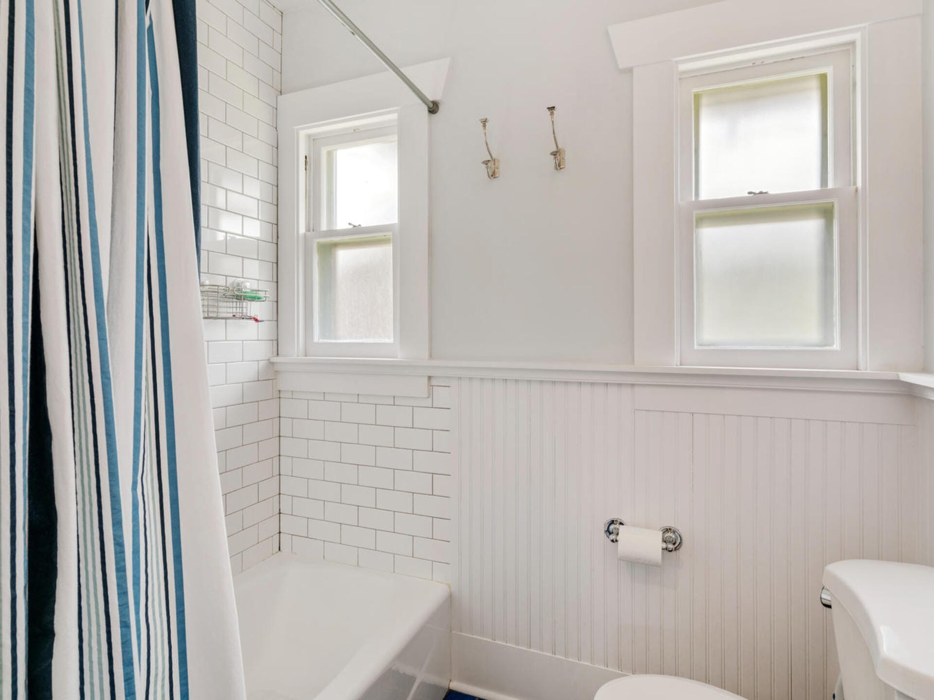 A bathroom with white subway tiles, a white tub, and a striped shower curtain. Two small frosted windows are above the tub. A white panel wall is below the windows, and a toilet paper holder is mounted next to the toilet.