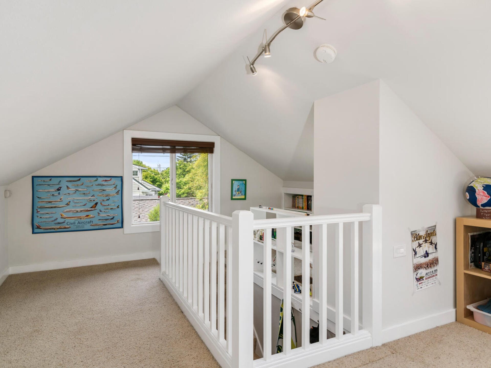 Loft-style room with slanted ceilings, featuring a window with a view of greenery outside. The space includes white railings, a carpeted floor, wall art, and shelves with books and a globe, creating a cozy reading nook. Ceiling track lighting above.