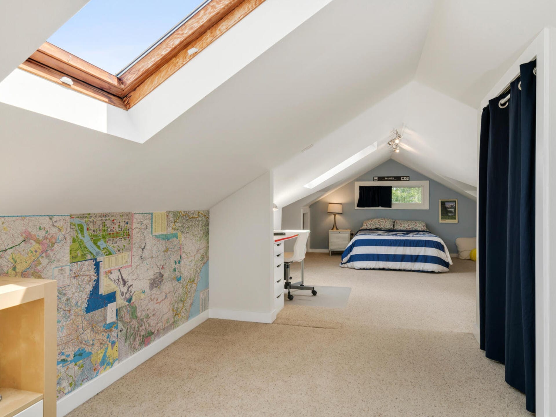 A cozy attic bedroom with slanted ceilings features a skylight, a map-covered wall, a small workspace with a desk and chair, and a bed with blue and white bedding. Natural light brightens the space, which is carpeted and decorated simply.