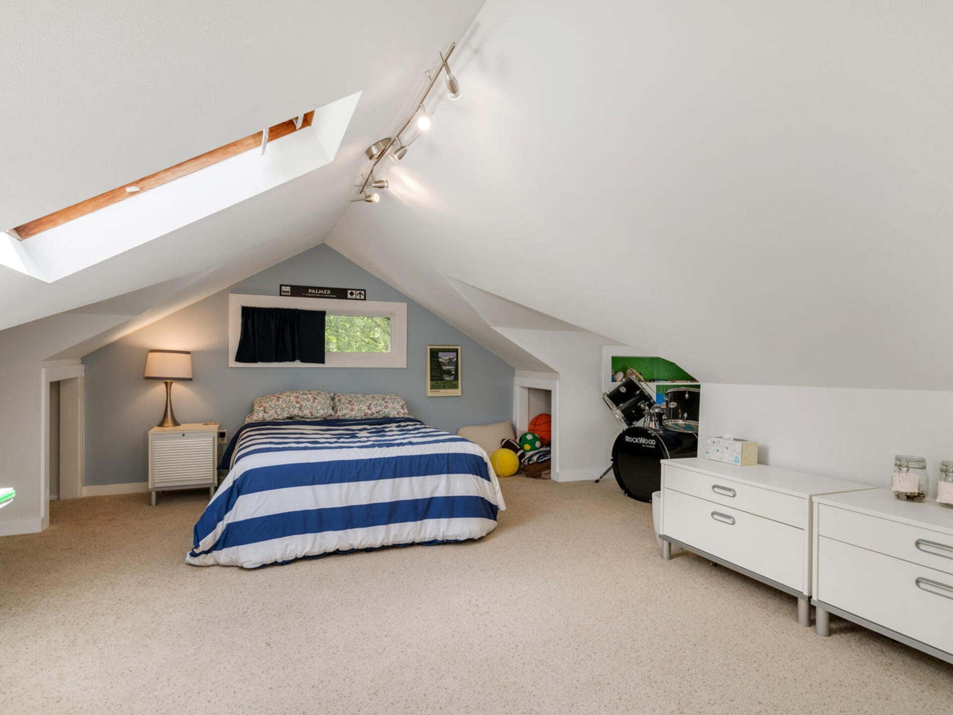 A cozy attic bedroom with a bed featuring blue and white striped bedding, placed under a skylight. On the right are a drum set and a white dresser. A small table lamp and framed pictures add warmth to the space.