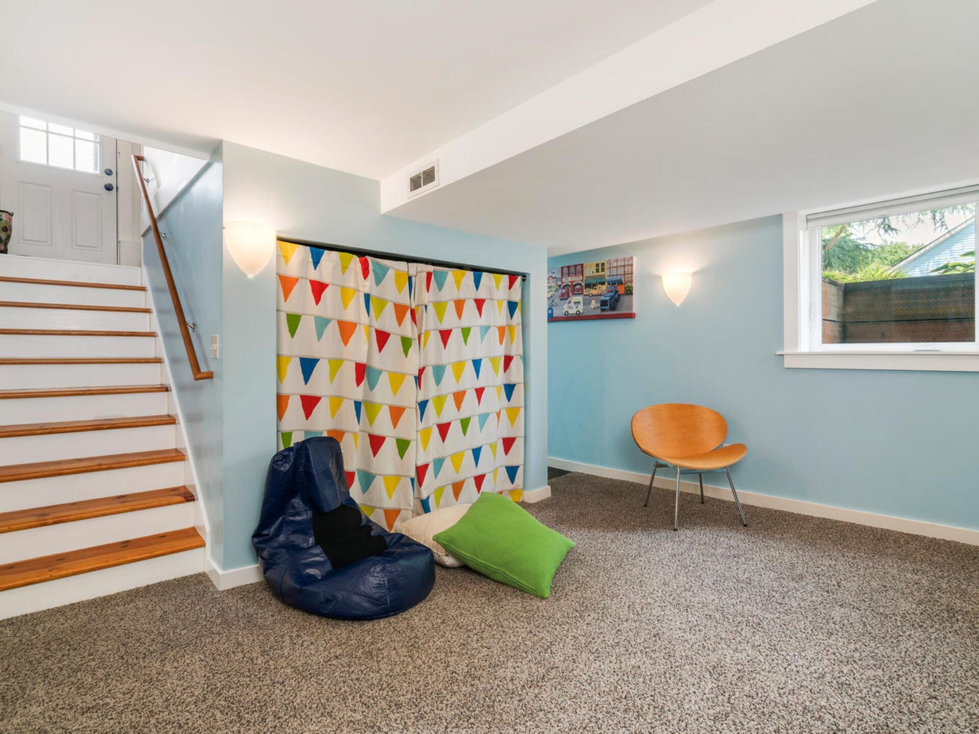 A cozy basement room with a bean bag chair, a colorful triangle-patterned curtain, and a wooden chair beside a window. Stairs with wooden steps lead to a small door. Walls are painted light blue, complementing the carpeted floor.